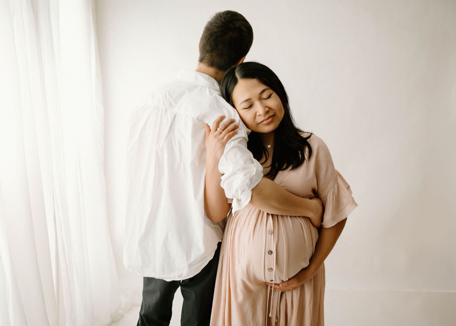 Couple maternity photo with pregnant woman in pink dress embracing partner before bright white studio backdrop.