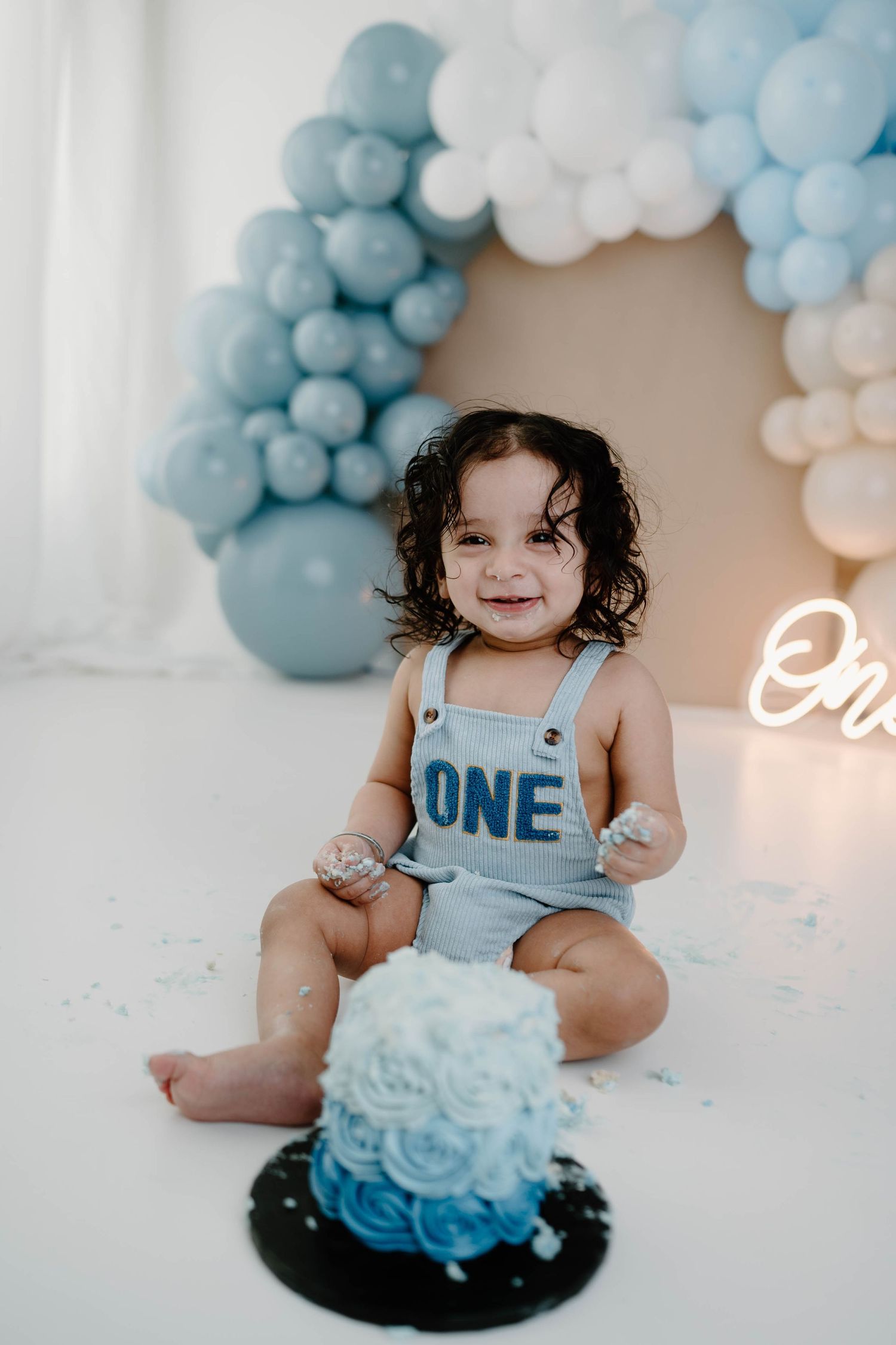 A happy toddler in a blue ONE romper smiles while sitting beside a blue ombre rosette smash cake.