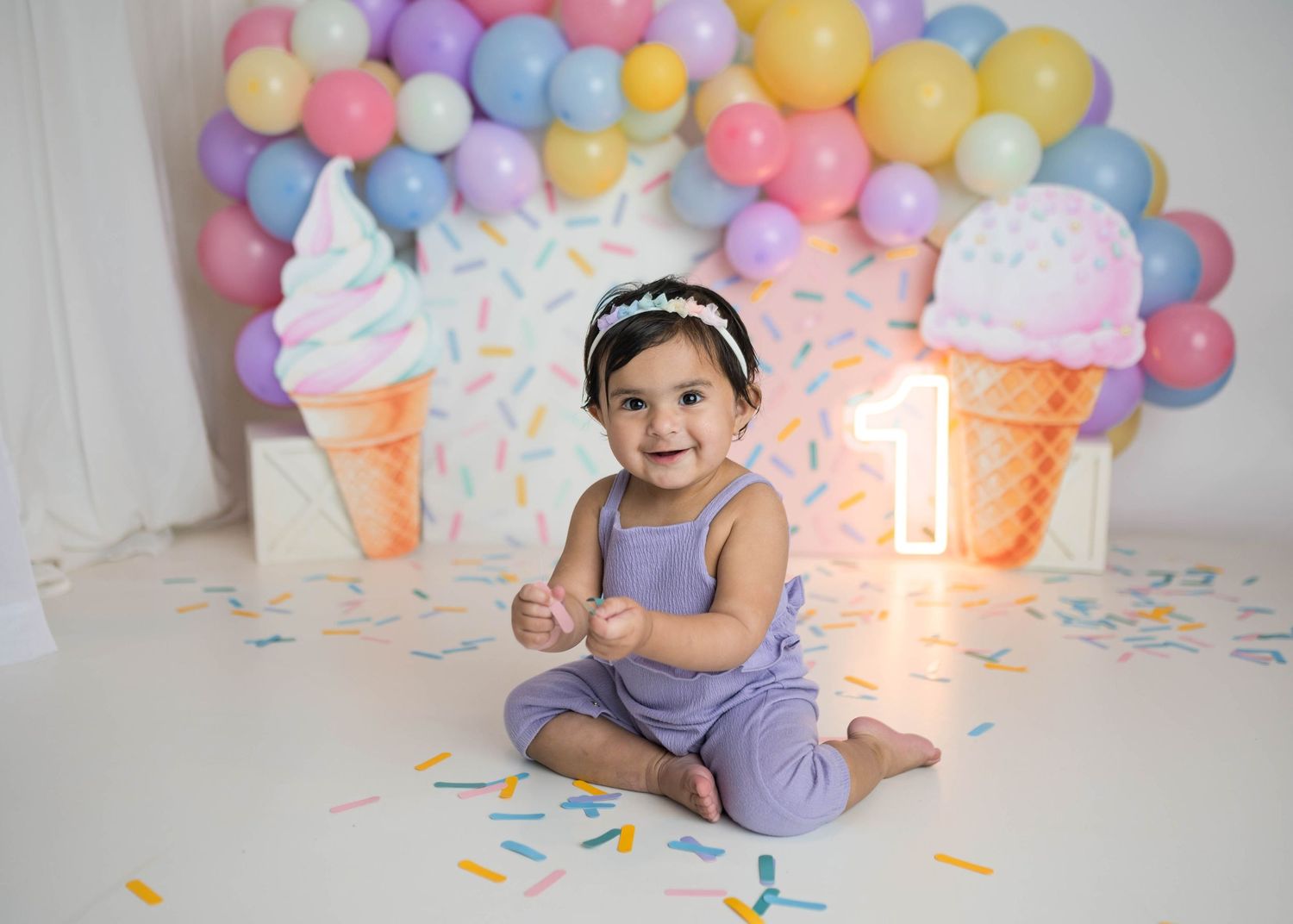 Cheerful baby in lavender romper sits on floor at ice cream themed first birthday with balloon backdrop.