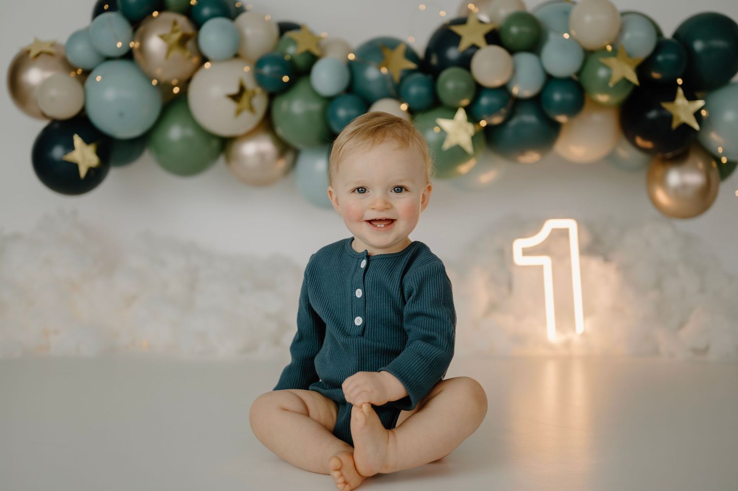 Happy baby boy in teal romper sitting before green gold balloon garland and glowing number 1.
