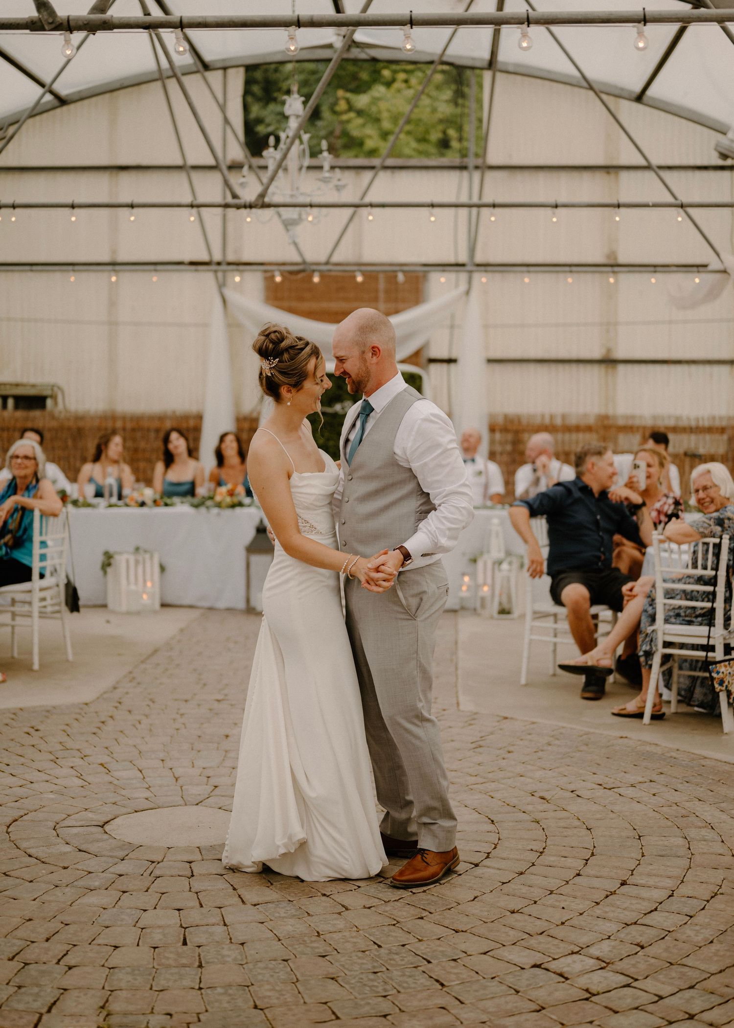 Bride and groom sharing first dance under string lights in a greenhouse wedding reception venue.