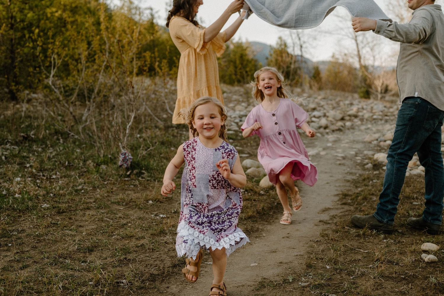 Two young girls running happily on a nature trail while parents play with a blanket behind them.