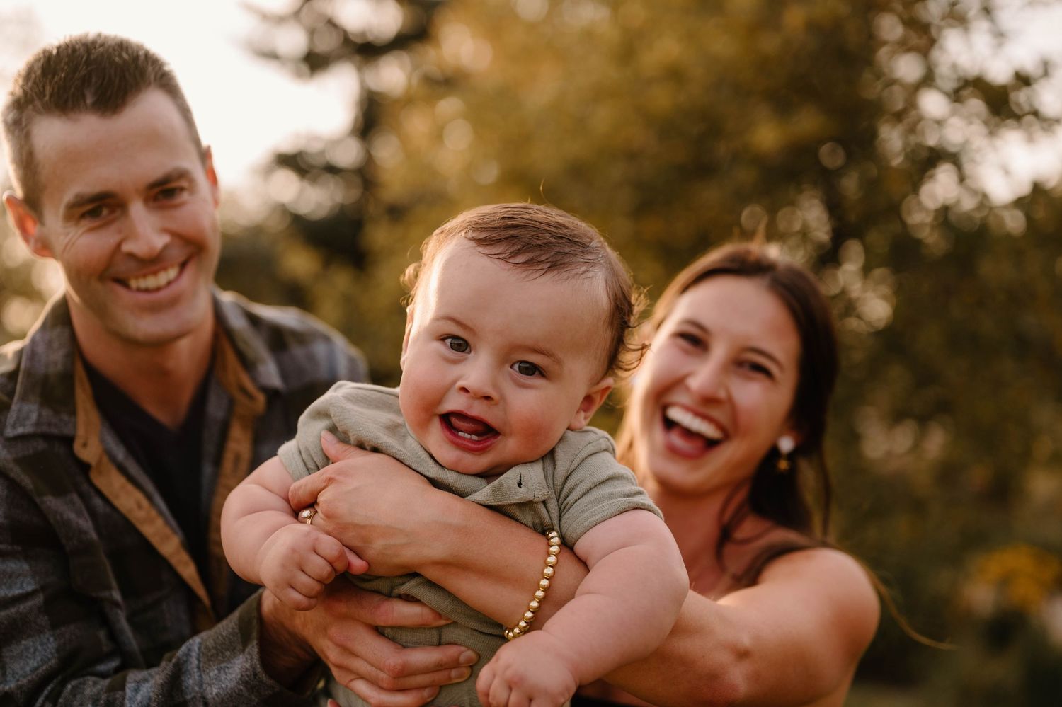 Happy family portrait with smiling parents holding cheerful baby boy in sunny outdoor setting.