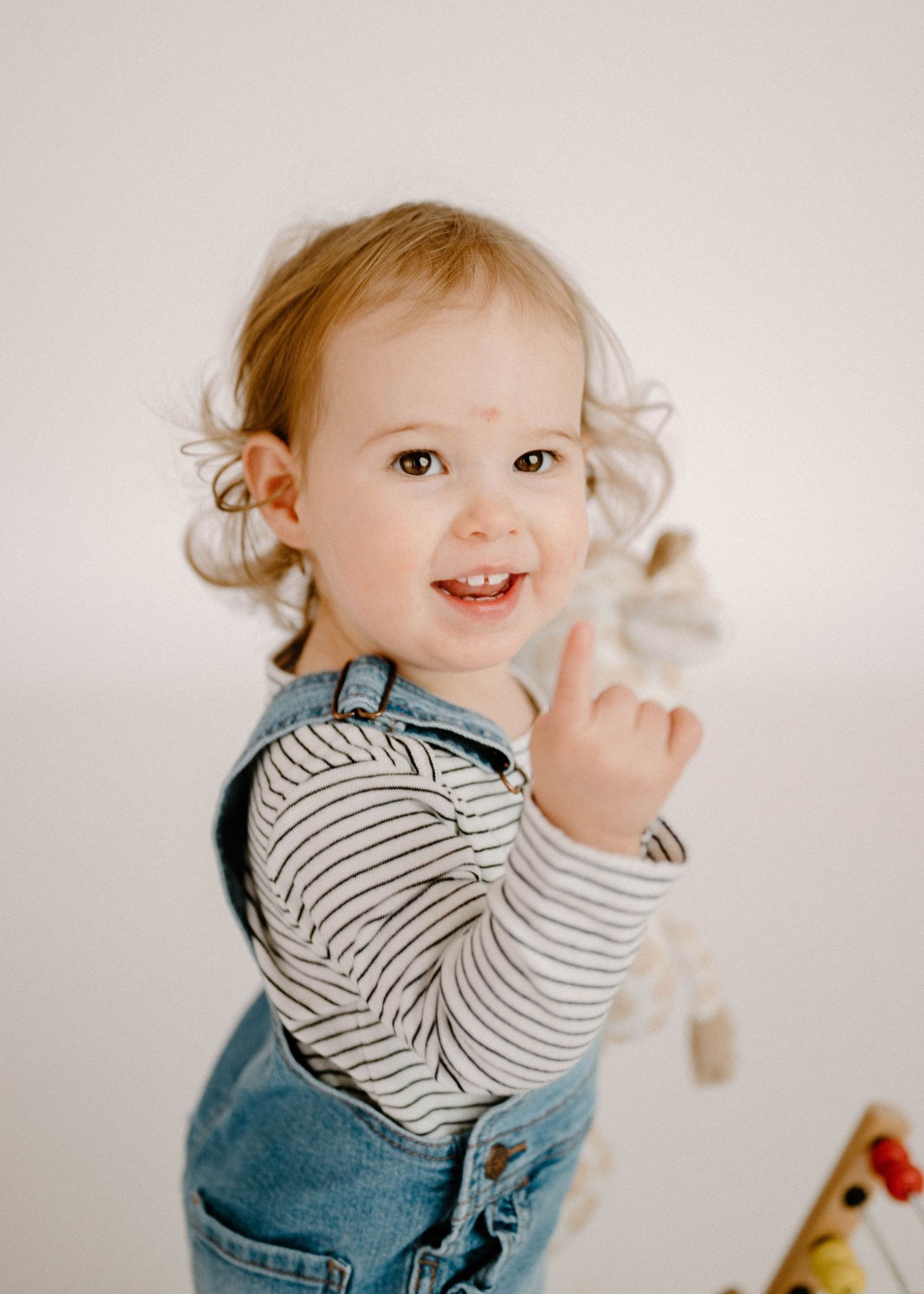 Cheerful toddler girl in denim overalls pointing finger upward and smiling at the camera.