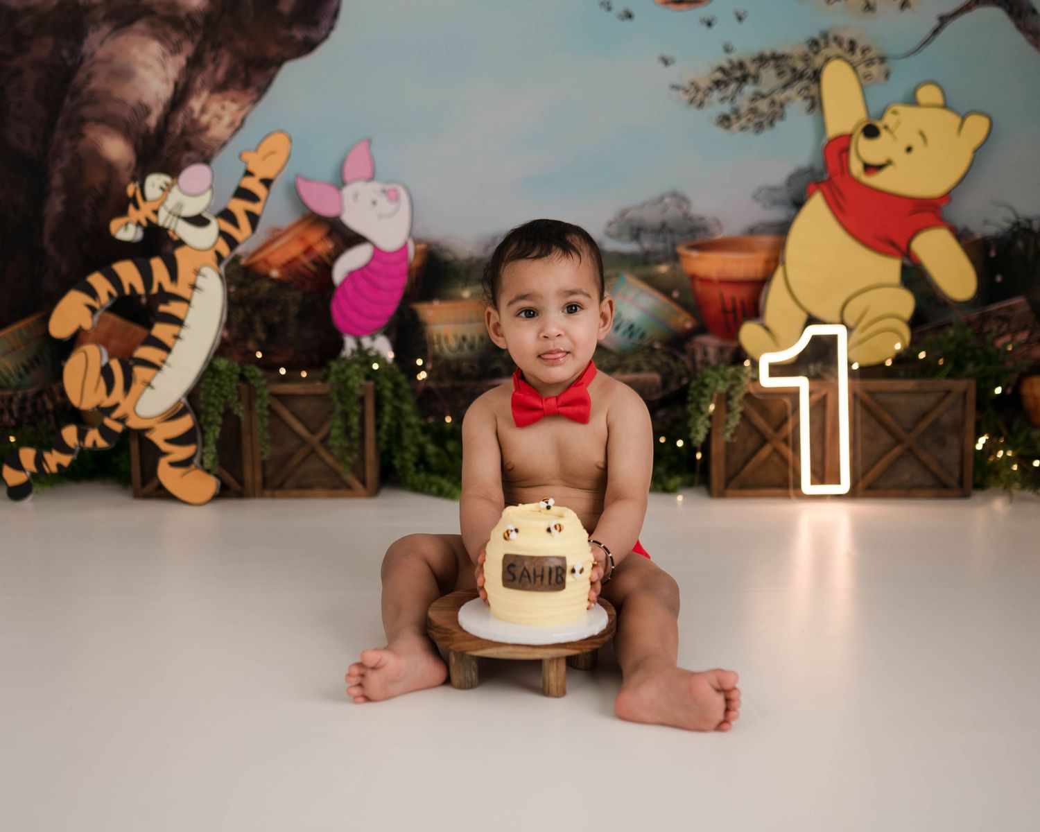 Baby boy in red bow tie sits with Winnie the Pooh themed cake and decorations celebrating his 1st birthday.
