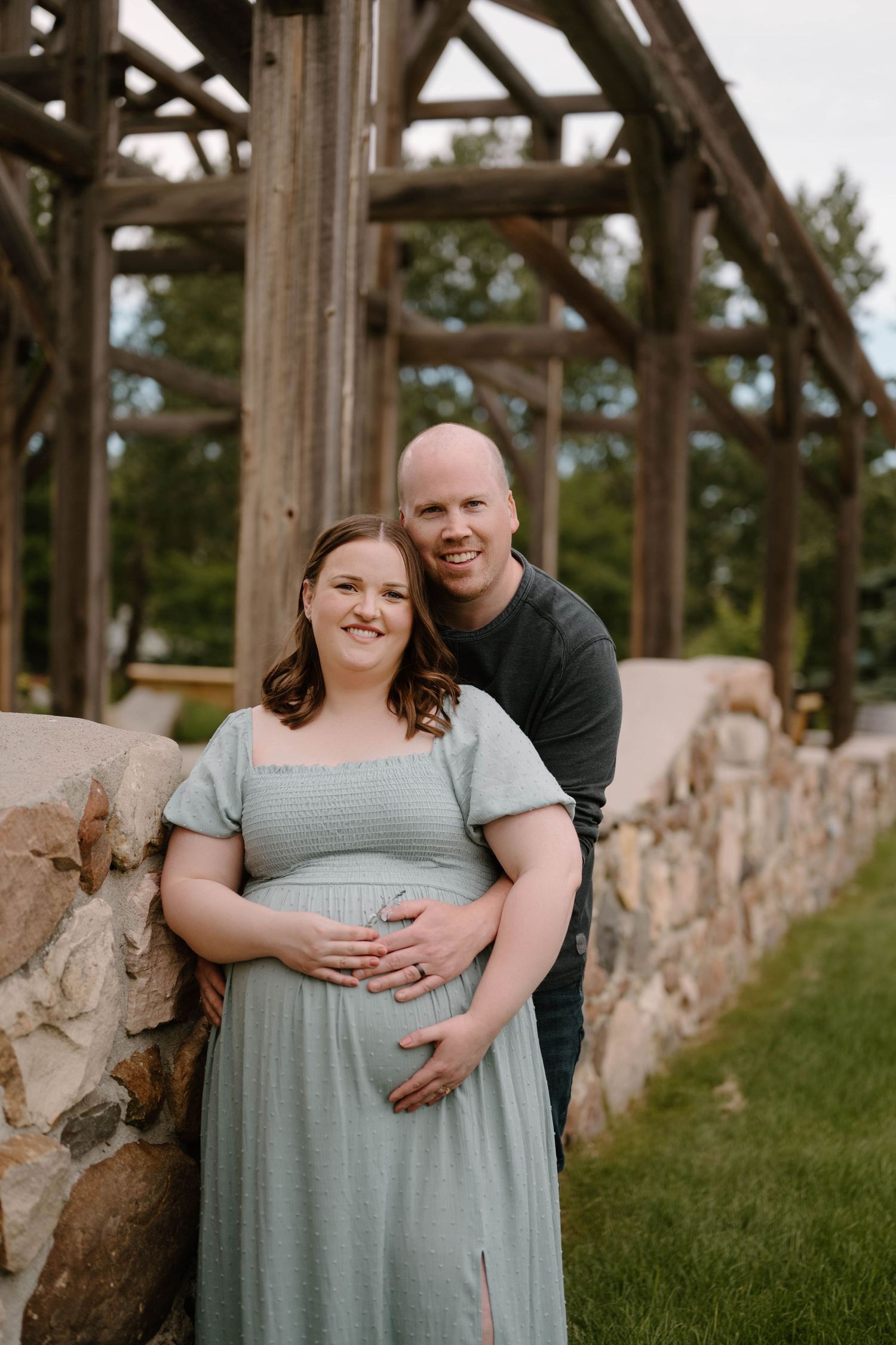 Smiling couple poses outdoors during maternity photo shoot, woman in sage green dress cradling baby bump near rustic stone wall.