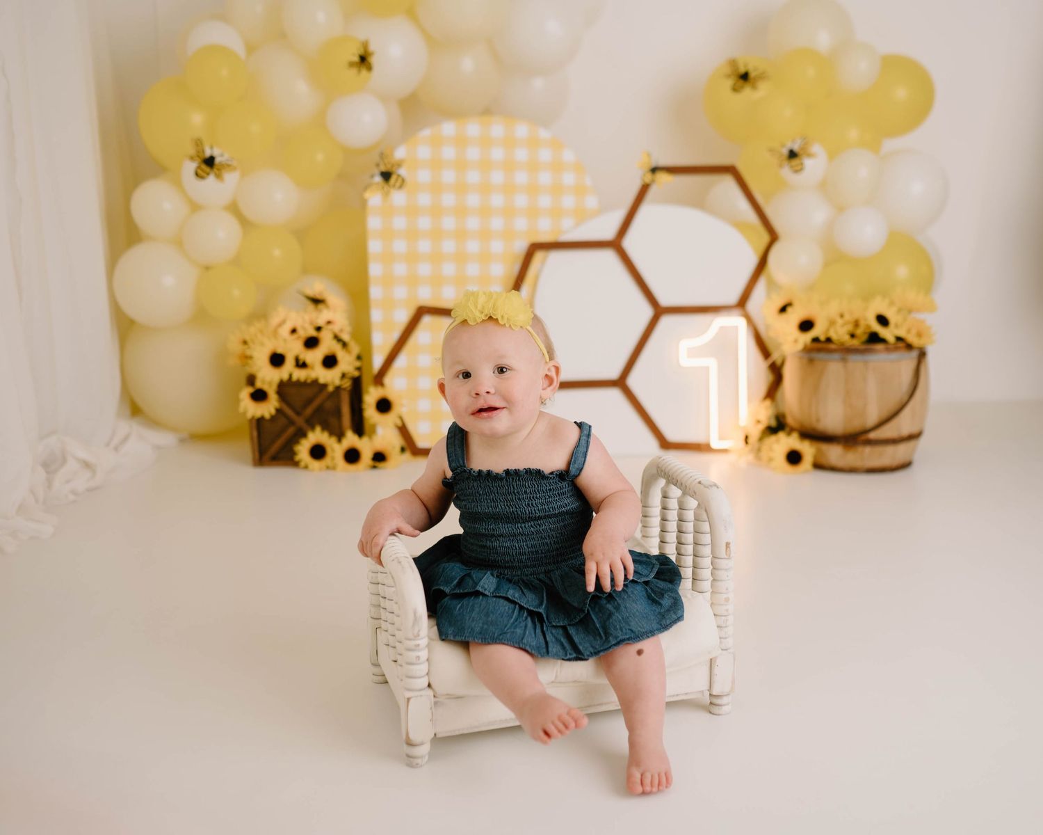 Baby girl in denim dress sits on white chair with bee-themed first birthday backdrop featuring honeycomb, balloons, and sunflowers.
