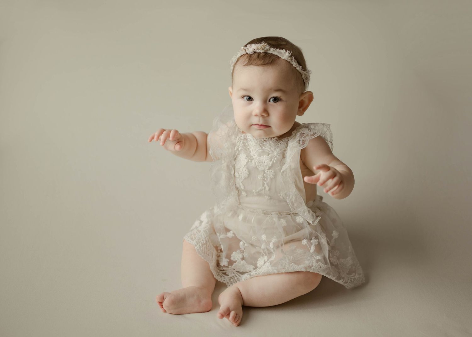 Cute baby girl in white lace dress and floral headband sitting on cream backdrop in studio milestone photo.