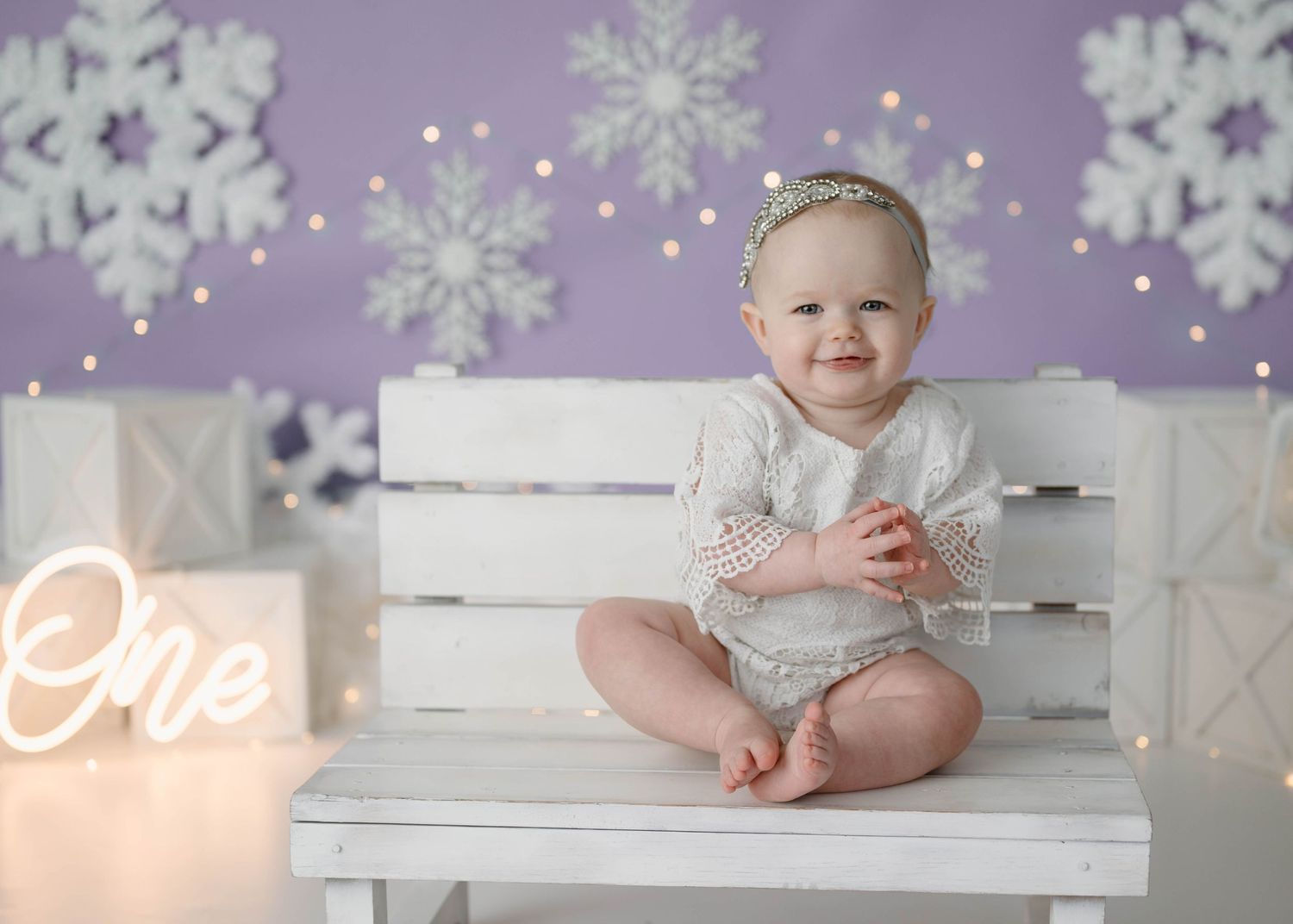 Smiling baby girl in white outfit sits on white bench with snowflake backdrop and One neon sign for first birthday.