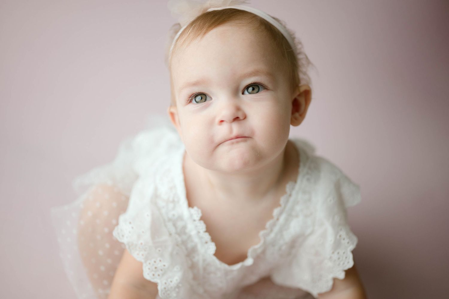Close-up portrait of baby girl in white lace dress with headband against soft pink studio background.