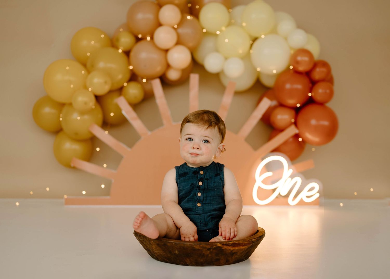 Baby boy in teal romper sitting in wooden bowl with boho sun balloon arch and One neon sign.