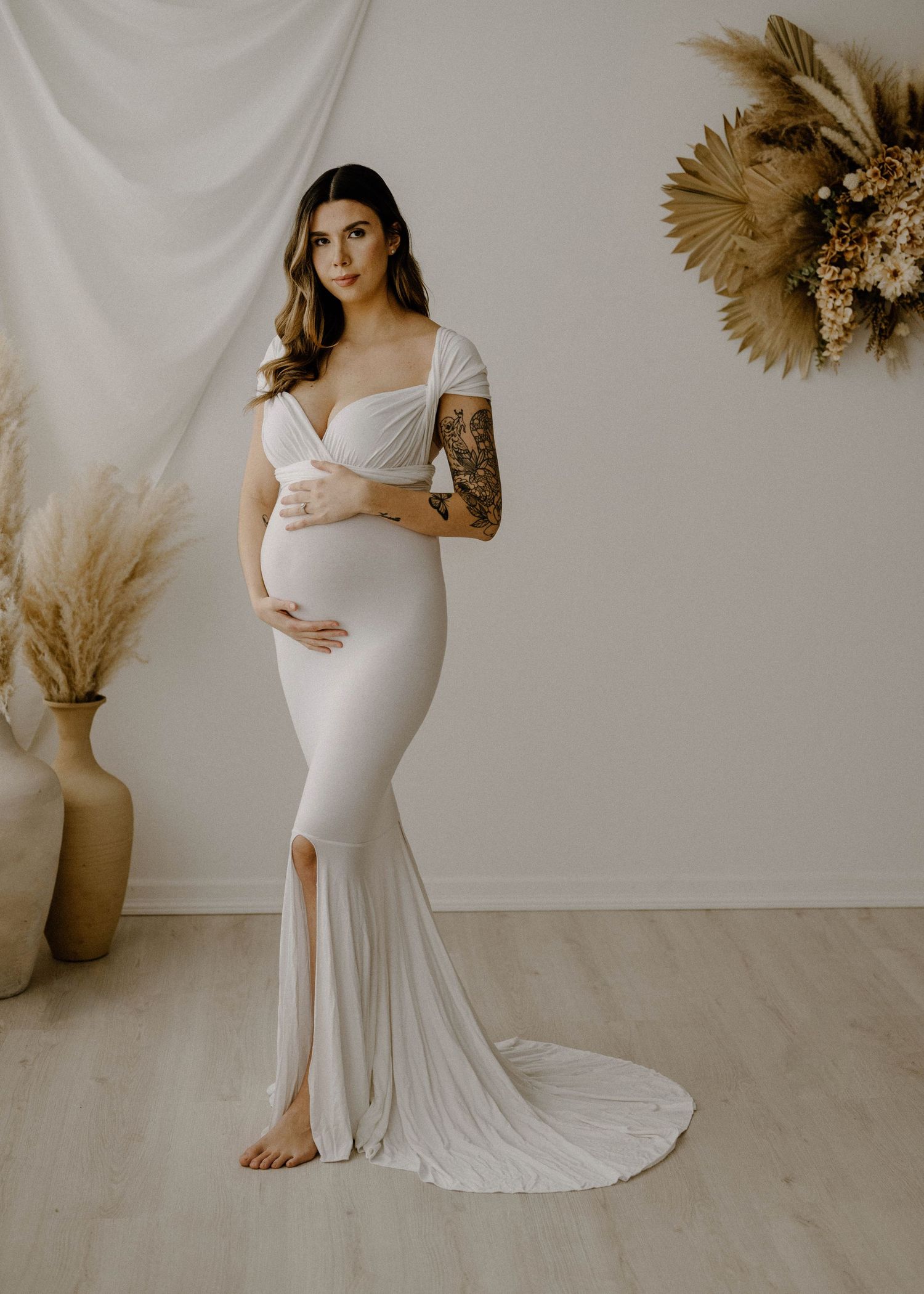 Pregnant woman in elegant white off-shoulder gown posing in a boho studio setting.