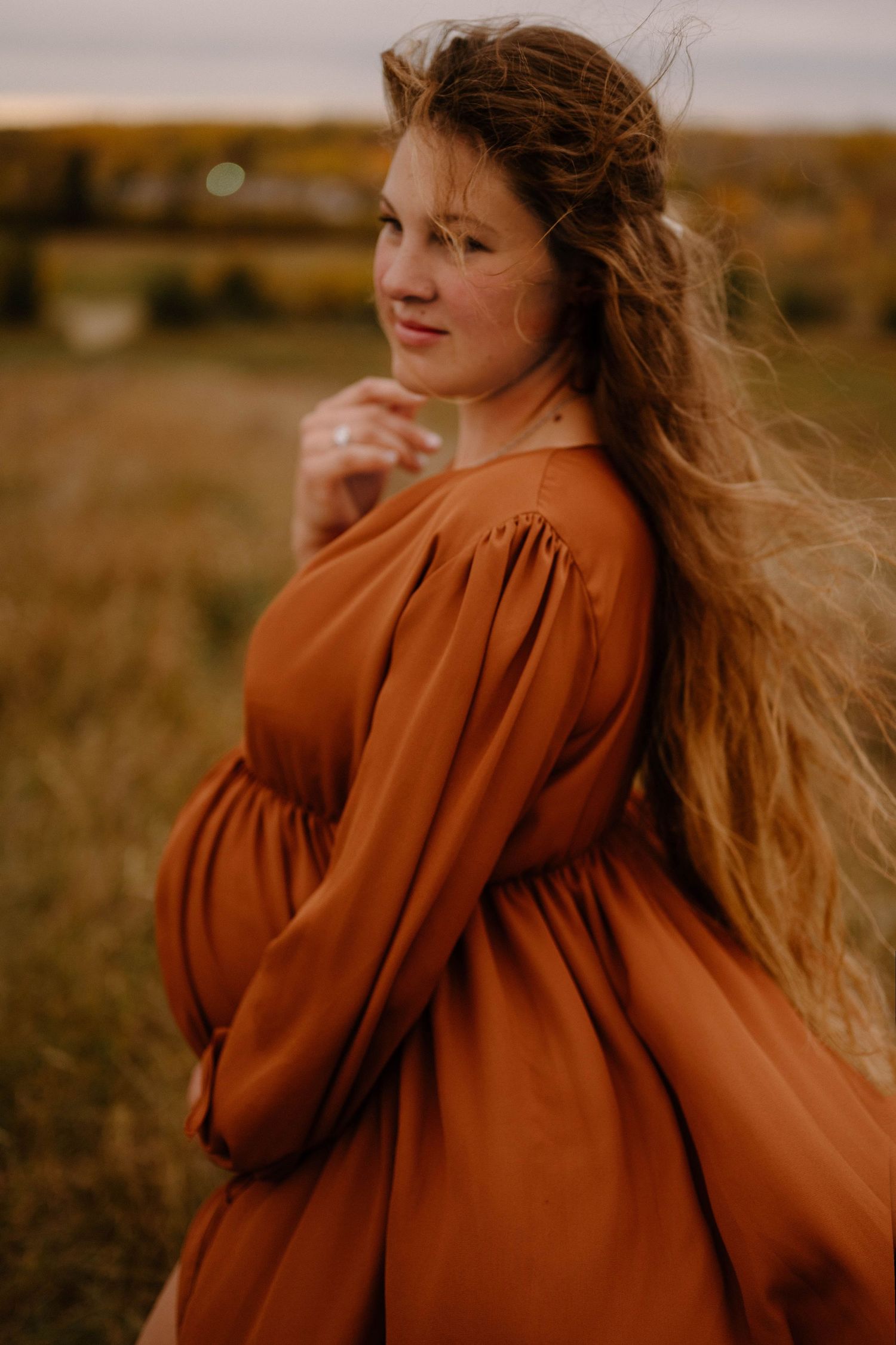 A smiling pregnant woman with windswept hair wears a rust-colored maternity dress in an autumn field.