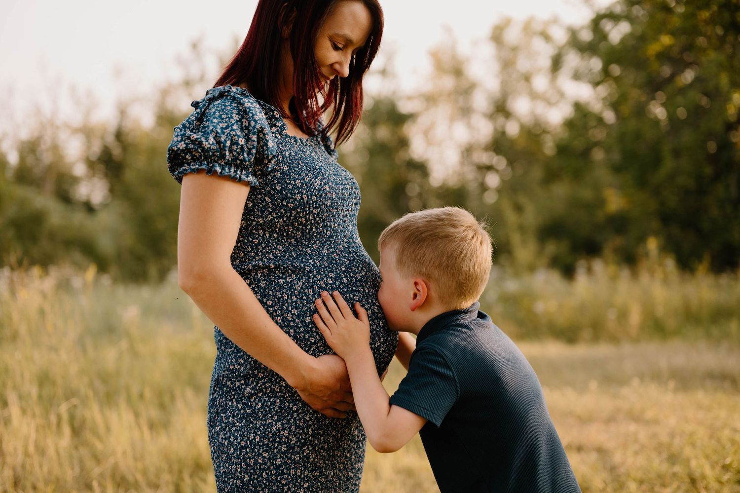 Young boy lovingly kisses pregnant mothers baby bump in a sun-drenched outdoor field at golden hour.