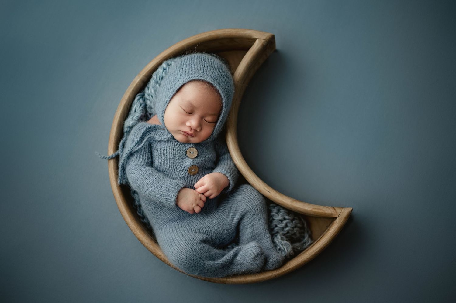 Newborn baby sleeping in knit blue outfit and bonnet in a wooden crescent moon prop.