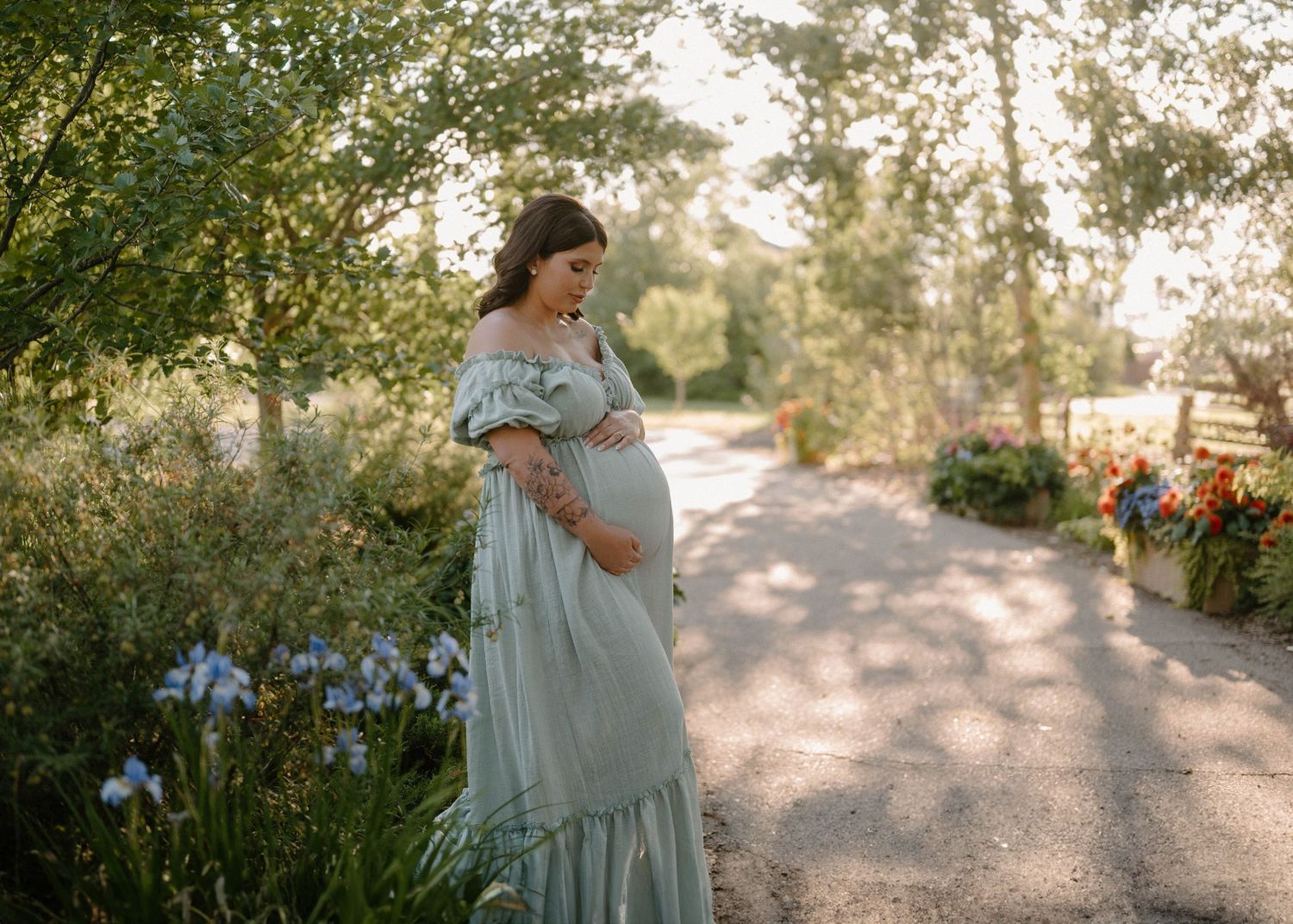 Pregnant woman in sage green off-shoulder gown standing on garden path surrounded by lush greenery and colorful flowers.