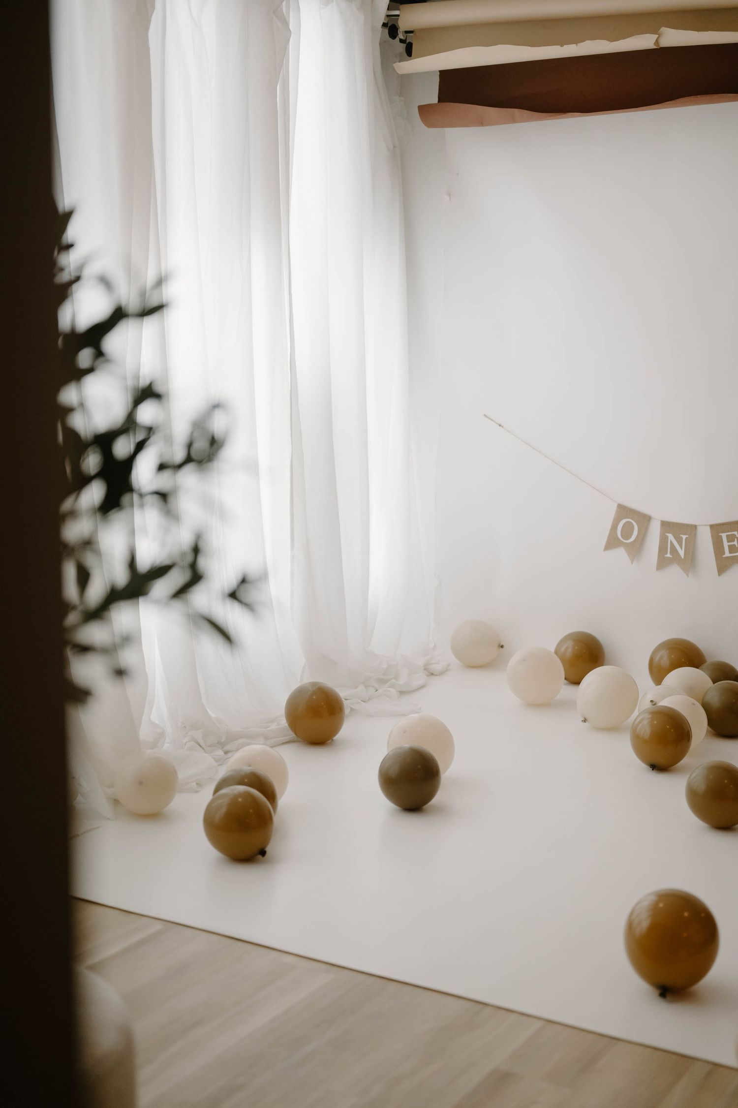 White and gold balloon first birthday party setup with ONE banner, sheer curtains, and scattered balloons on white backdrop.