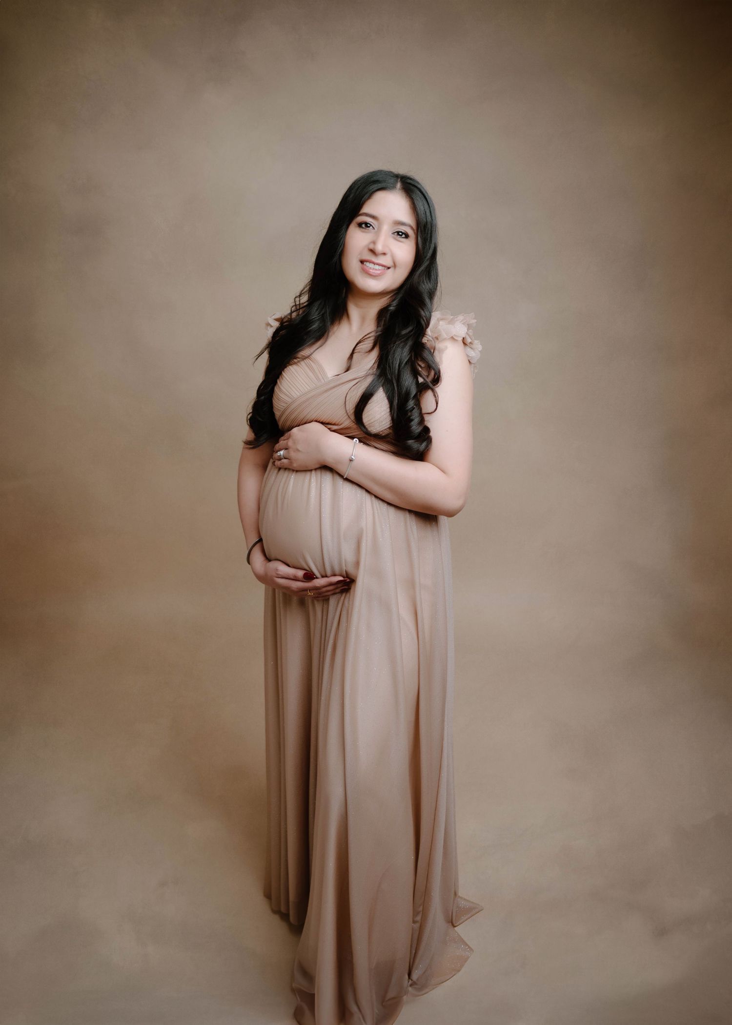 A pregnant woman in a flowing beige dress cradles her baby bump in a professional maternity portrait.