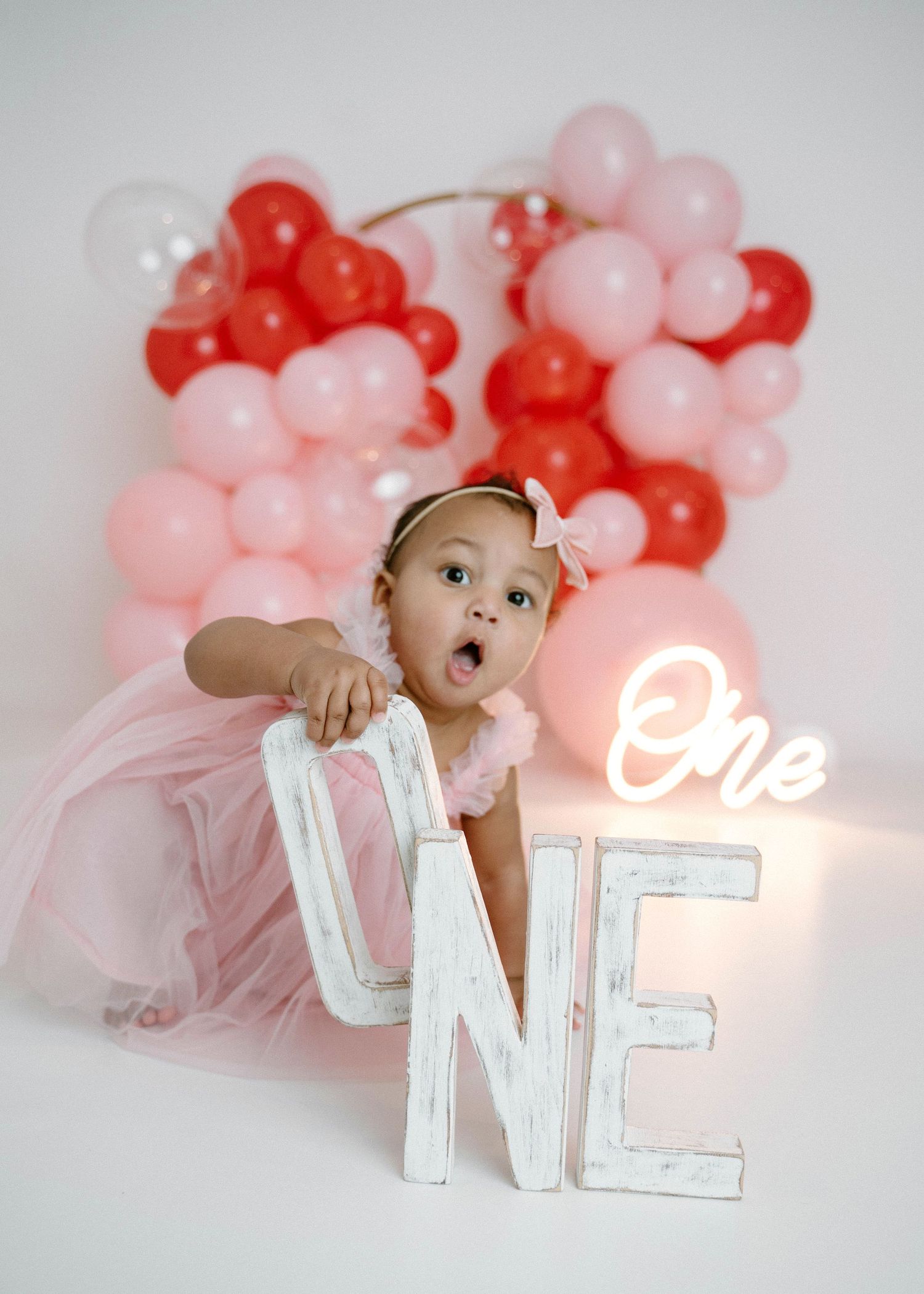 Baby girl in pink tulle dress peeks over ONE letter sign with colorful balloon backdrop behind.