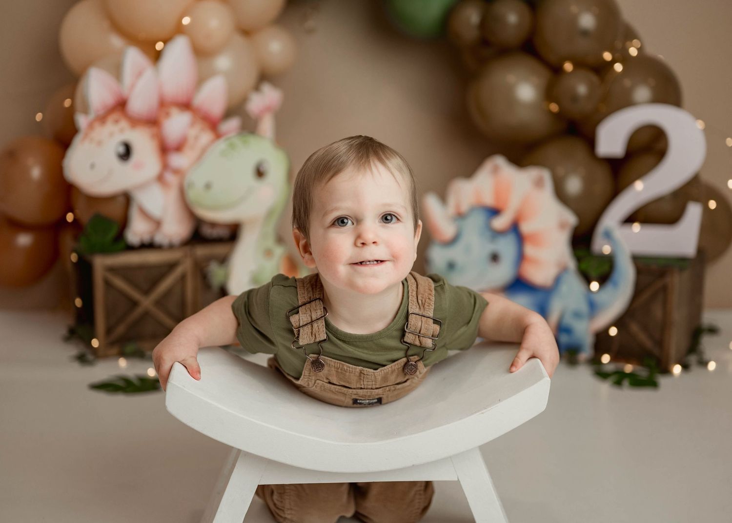 Toddler boy in green shirt leans on stool at dinosaur second birthday cake smash session.