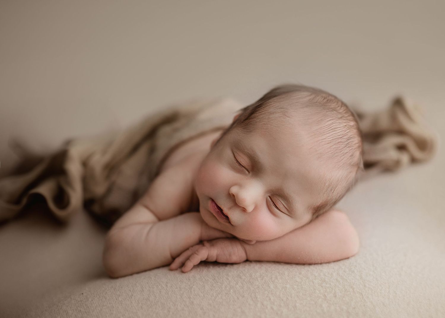 Peaceful smiling newborn baby resting chin on hands with brown textured wrap on cream surface.