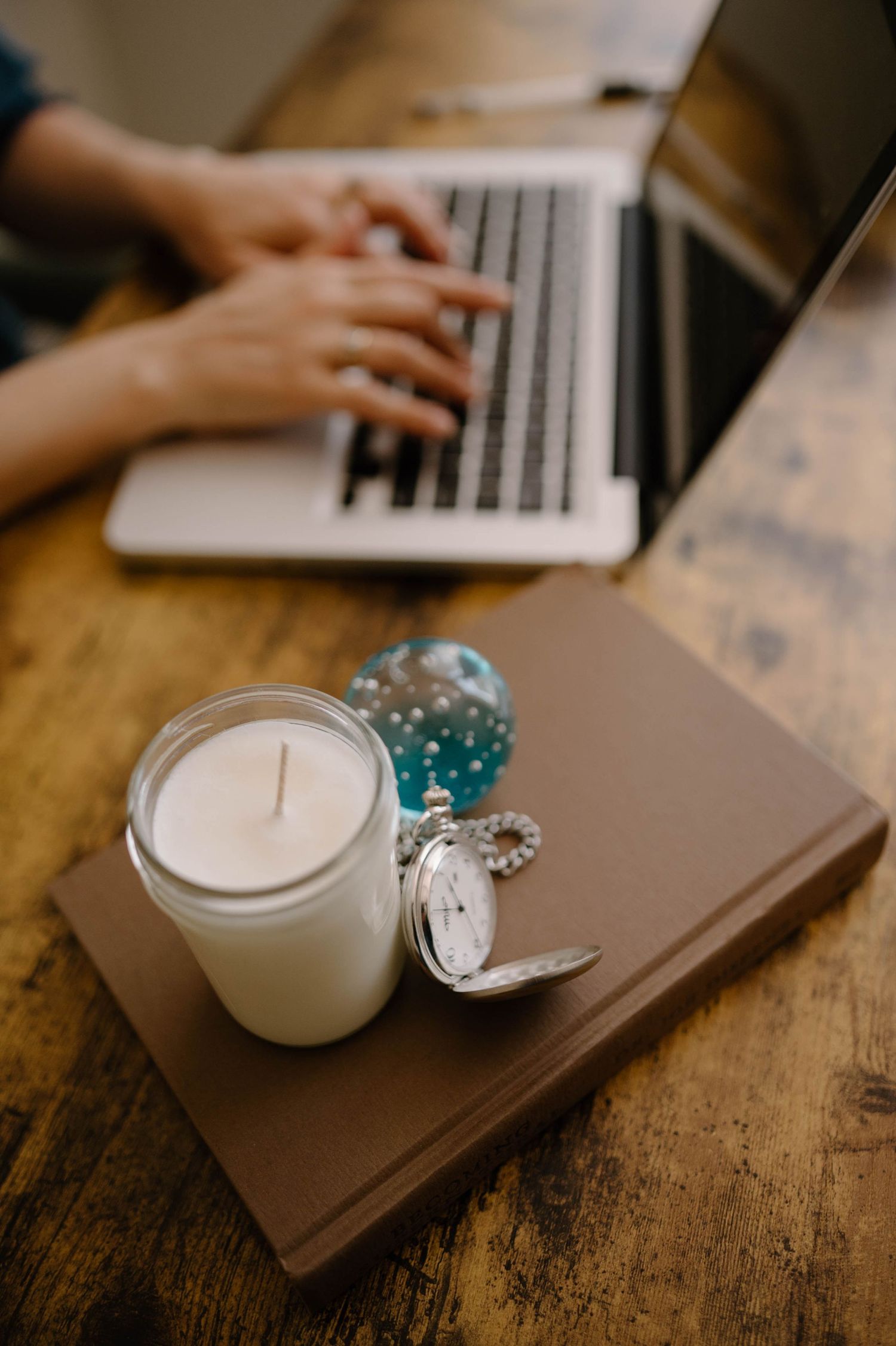 Close-up of candle, pocket watch, and book on wooden desk with person typing on laptop in background.