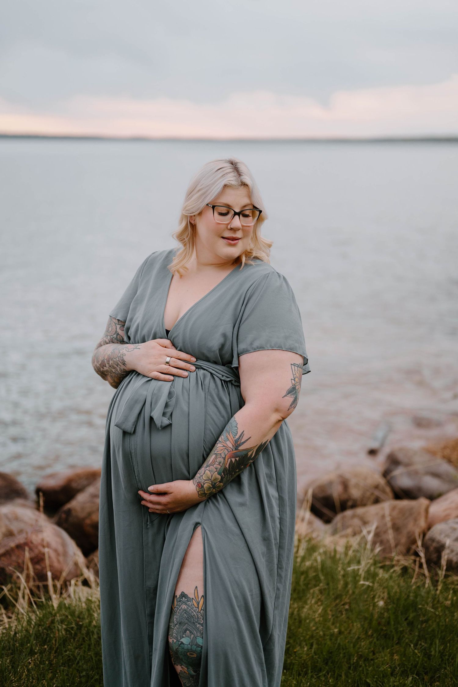 Pregnant woman in sage green maxi dress with tattoos stands by rocky shoreline, cradling her baby bump at sunset.
