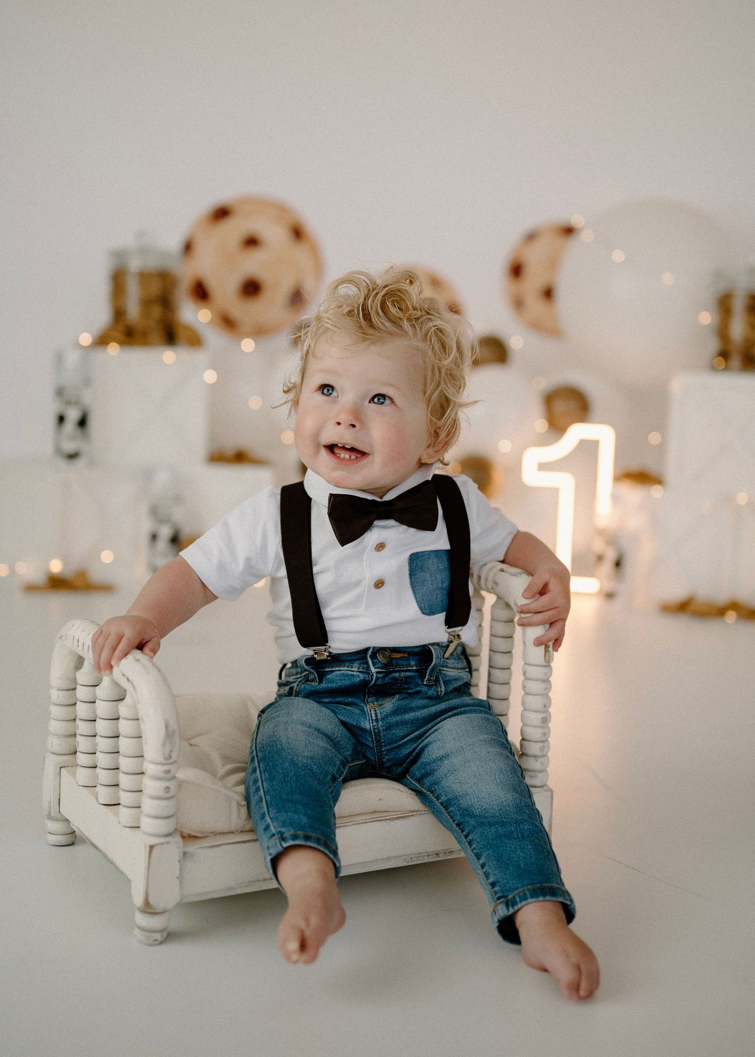 Curly-haired toddler in suspenders sitting on white mini bed with cookies and milk birthday theme.