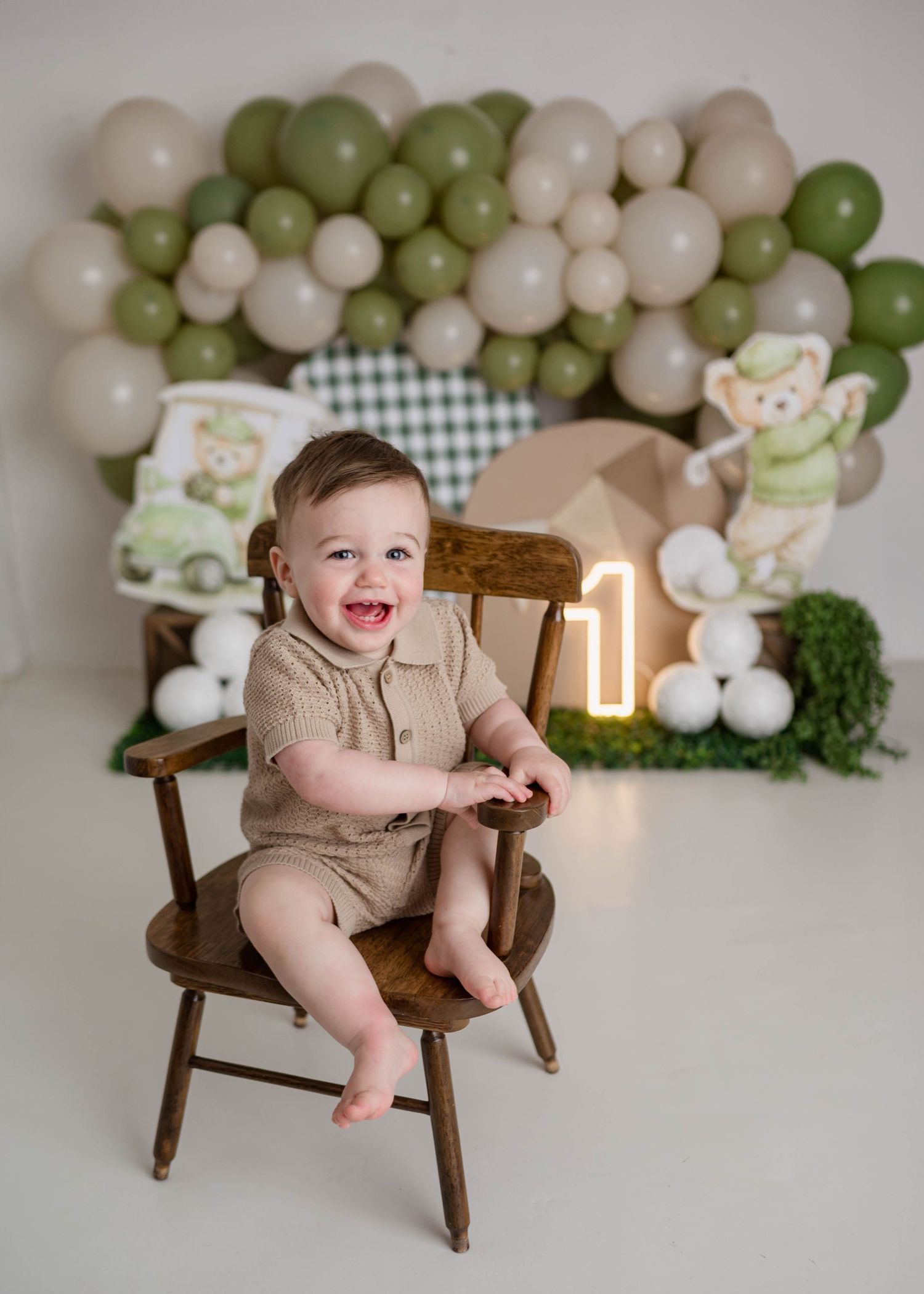 A smiling baby boy sits on a wooden chair at his first birthday party with green balloons and a glowing number one.