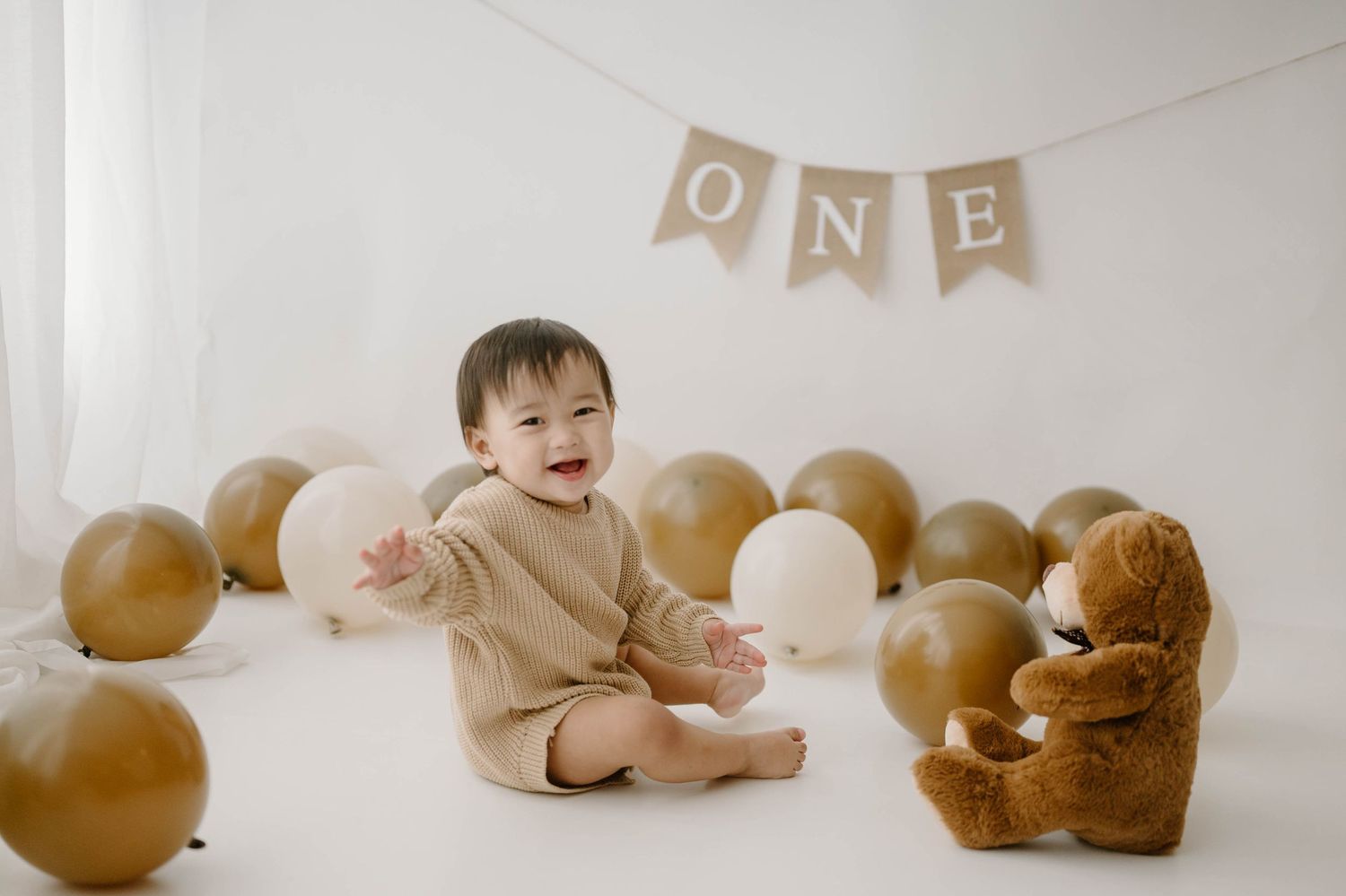 Happy baby celebrates first birthday sitting among gold and white balloons with a teddy bear and ONE banner.