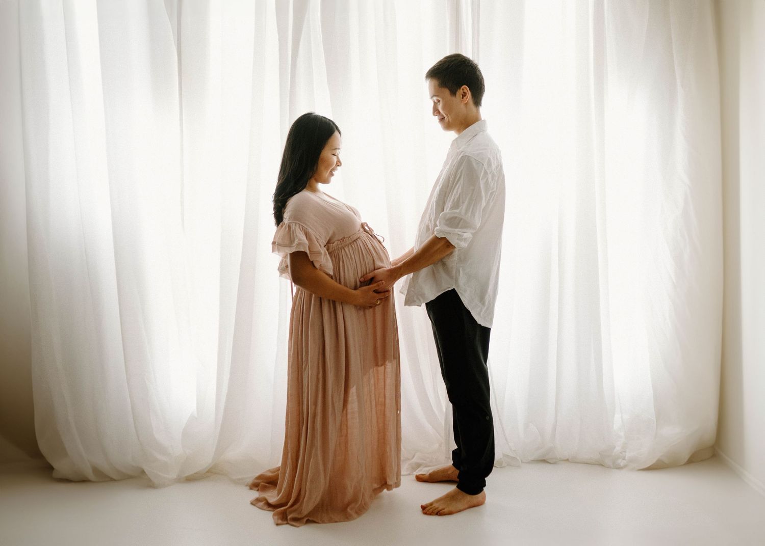 Expectant couple holding hands and gazing at each other in front of sheer white curtains.