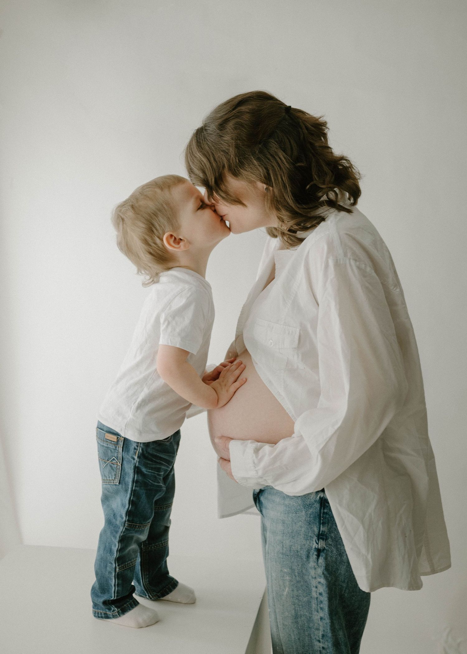 Pregnant woman and young toddler share a sweet nose-to-nose kiss in a bright white studio.