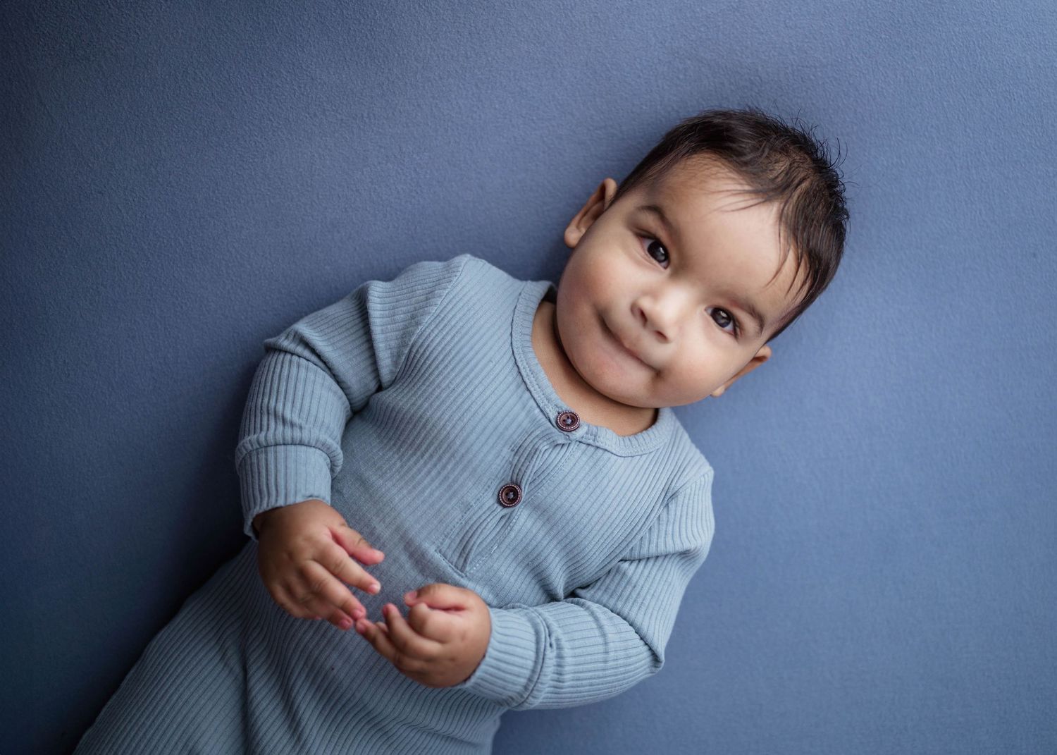 Adorable baby in a blue ribbed romper lying on a blue backdrop, looking at the camera with a gentle smile.