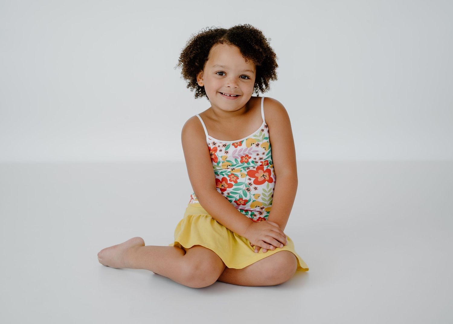 Smiling young girl with curly hair wearing a floral top and yellow skirt, sitting on a white studio floor.