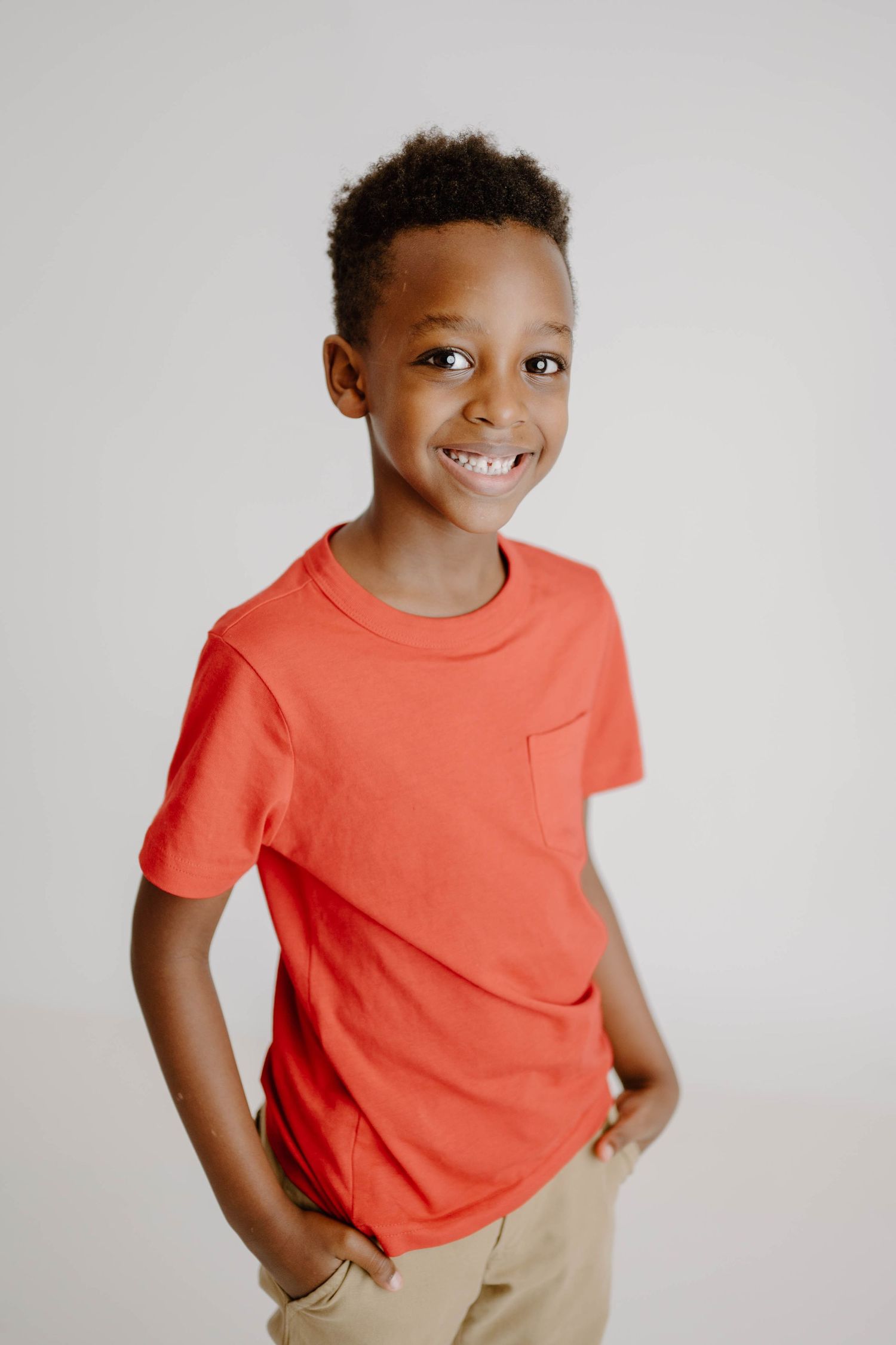 Smiling young Black boy in a red pocket t-shirt and khaki shorts posing with hands in pockets.