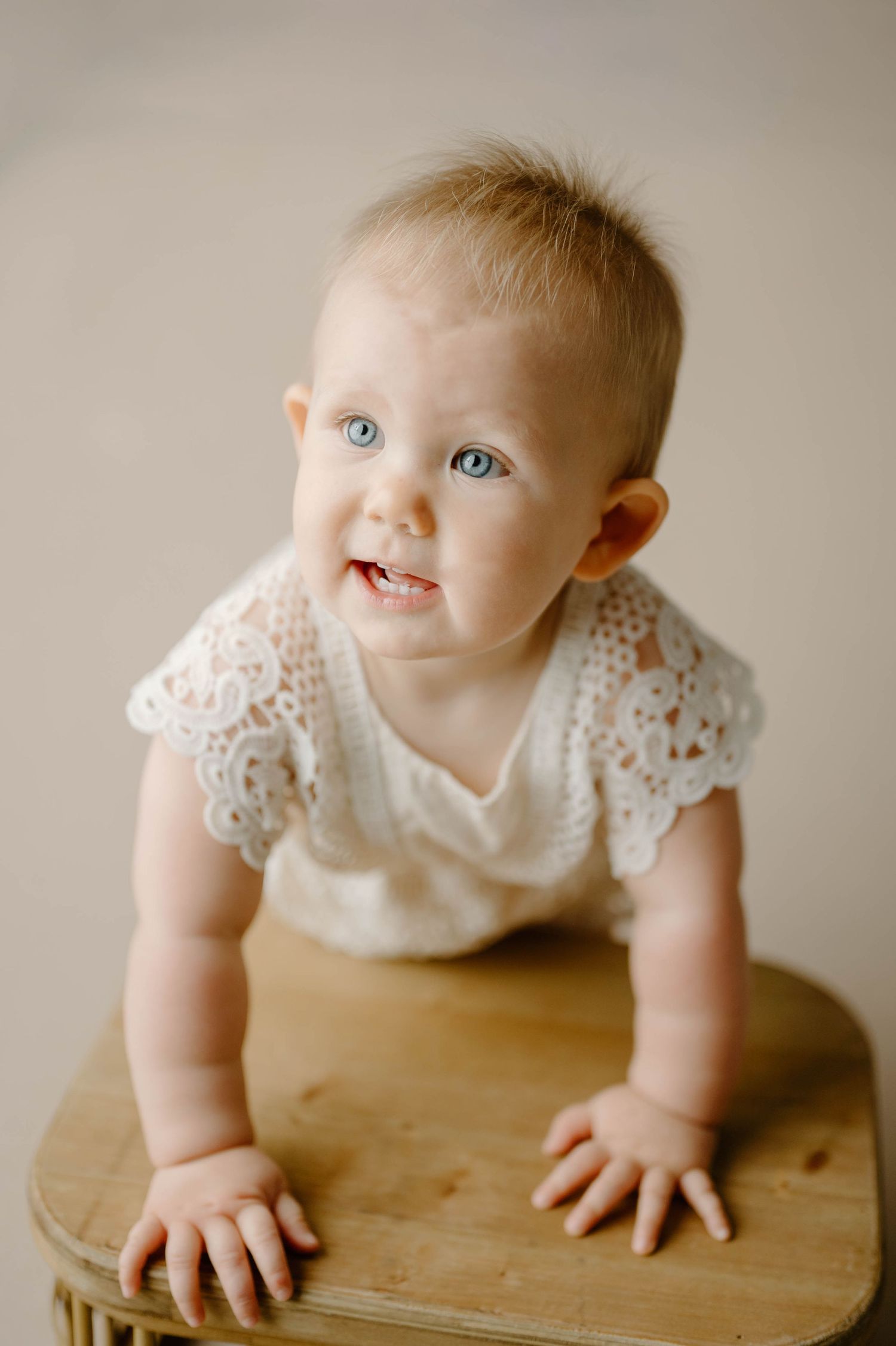 Blue-eyed baby girl in white lace romper leaning on a wooden stool during a studio photography session.