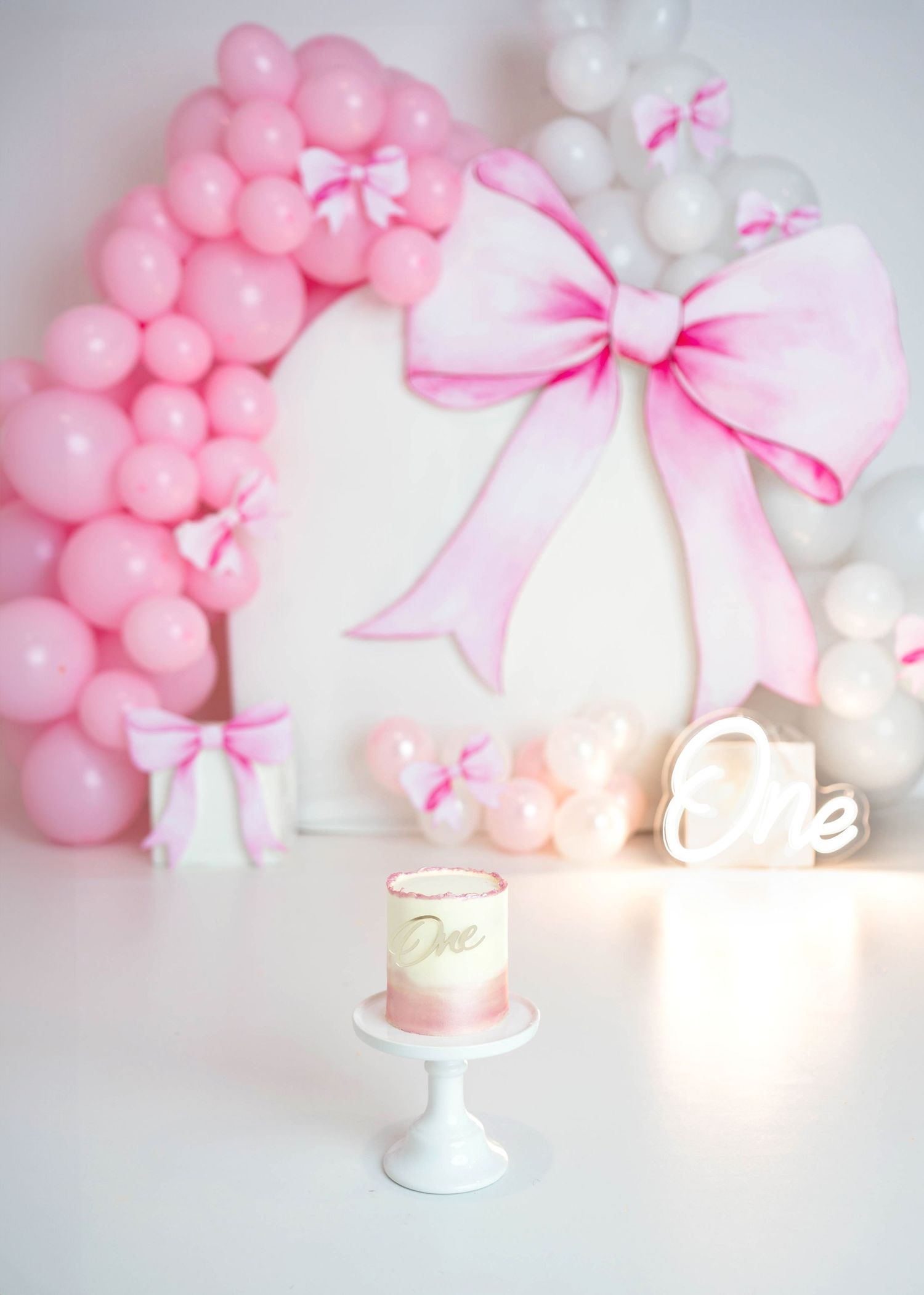 Pink and white first birthday cake smash setup with bow decorations and balloon garland.