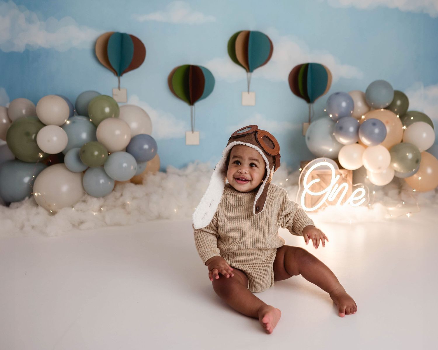 Happy baby wearing a vintage aviator hat sitting on a white floor at a hot air balloon themed cake smash session.