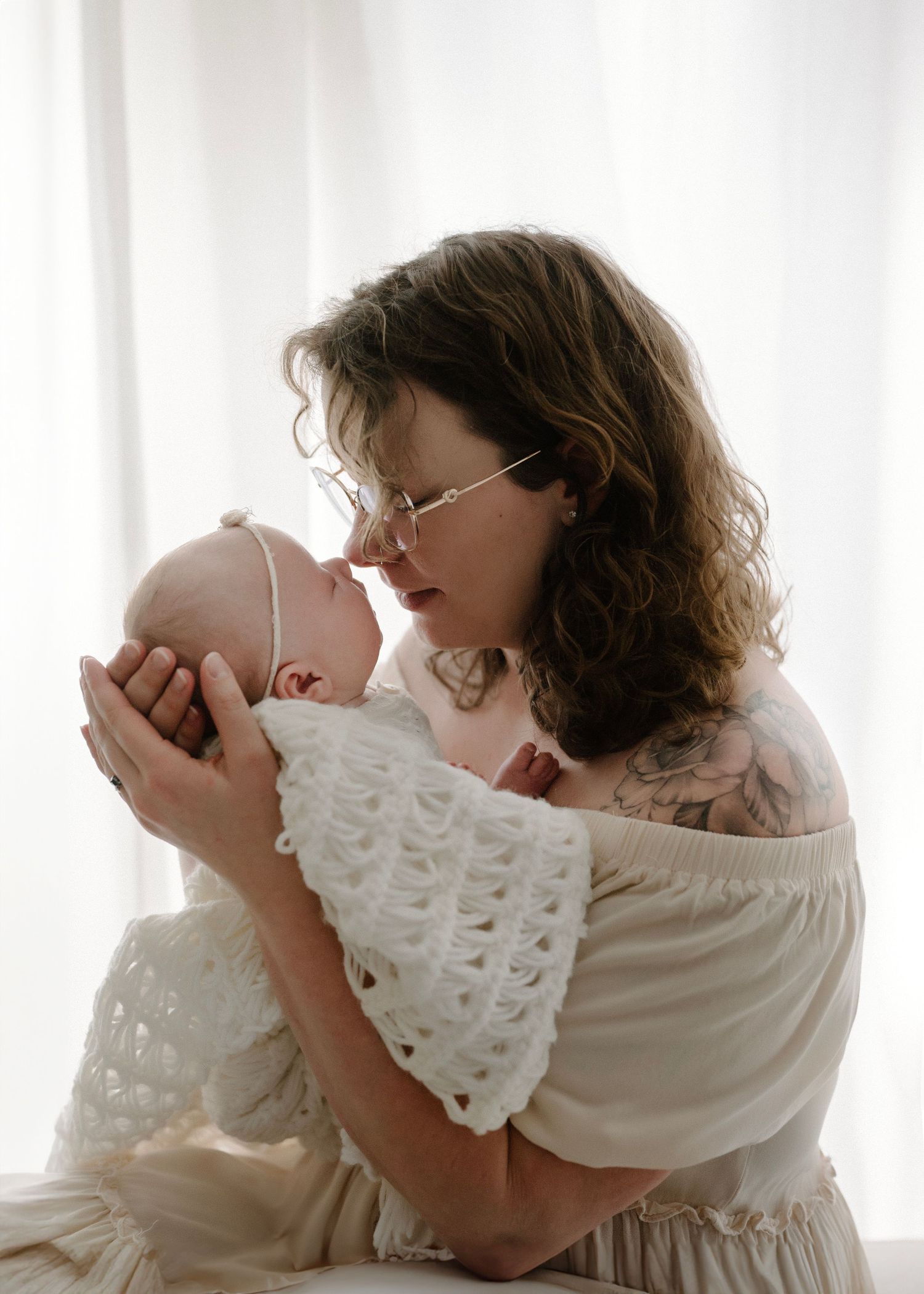 A tattooed mother with curly hair and glasses tenderly holds her newborn baby in a white crocheted wrap.
