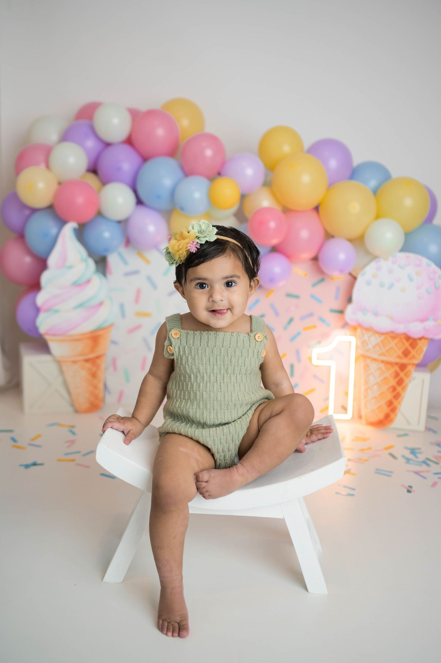 Baby girl in green romper sits on white chair at ice cream themed first birthday with colorful balloon backdrop.