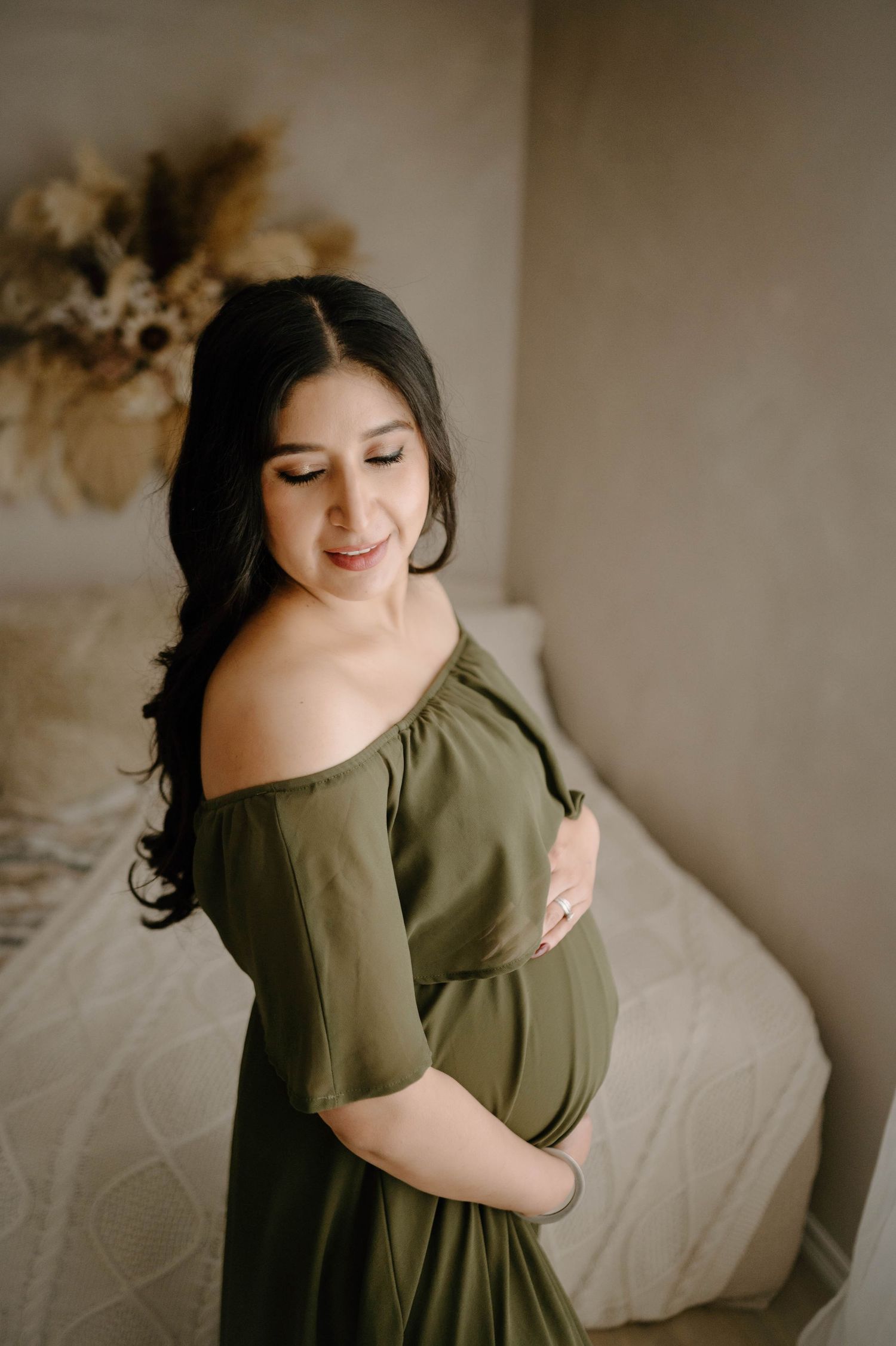 Smiling pregnant woman in olive maternity dress cradling baby bump near bedroom setting.