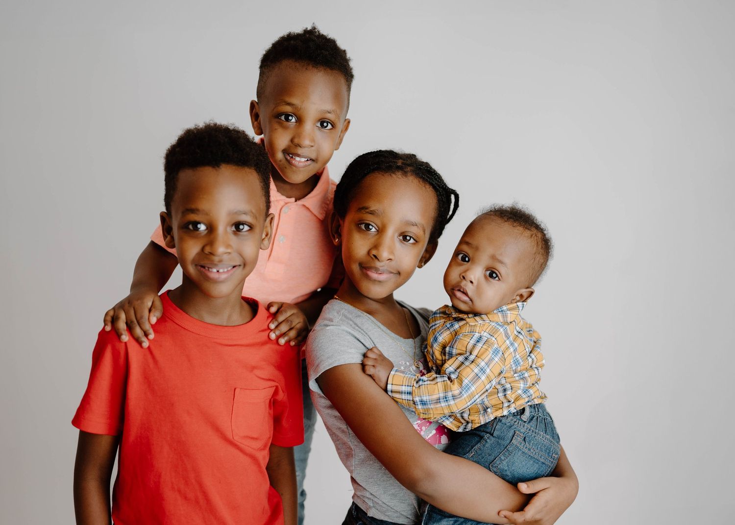 Four young siblings posing together in a studio, with the oldest holding the baby in a warm family portrait.