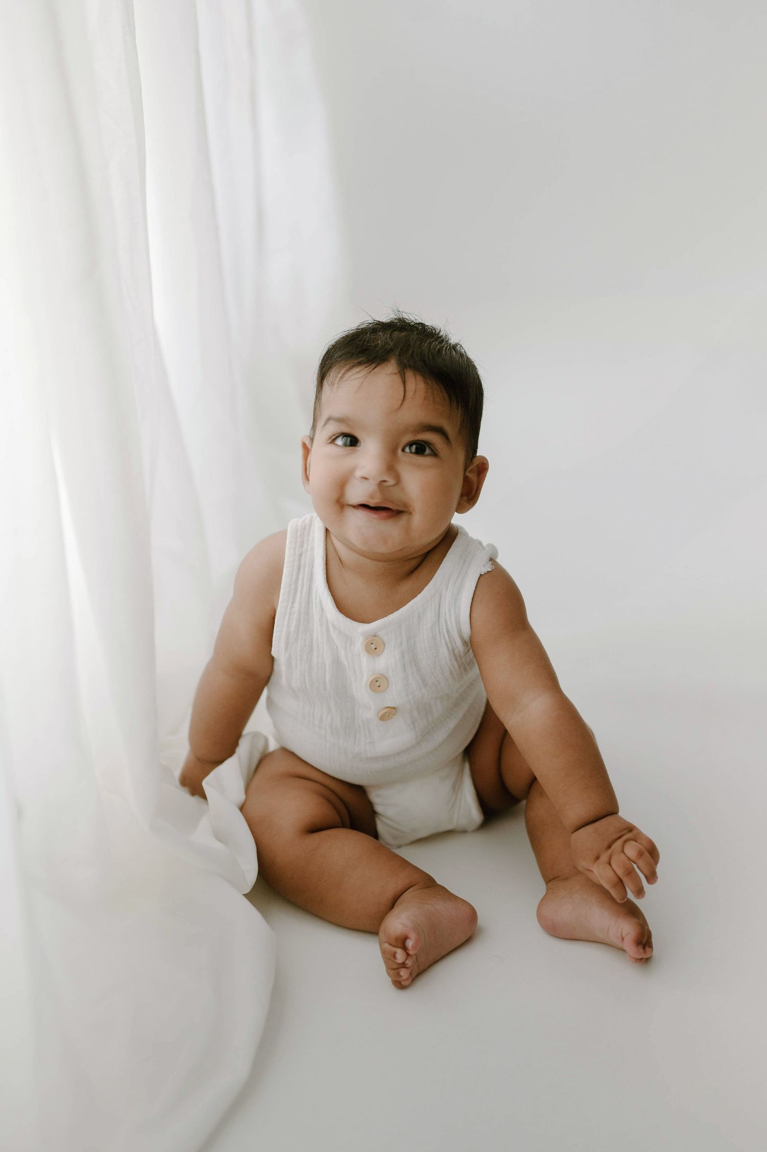 Cheerful baby in white romper sitting near white curtain smiling.
