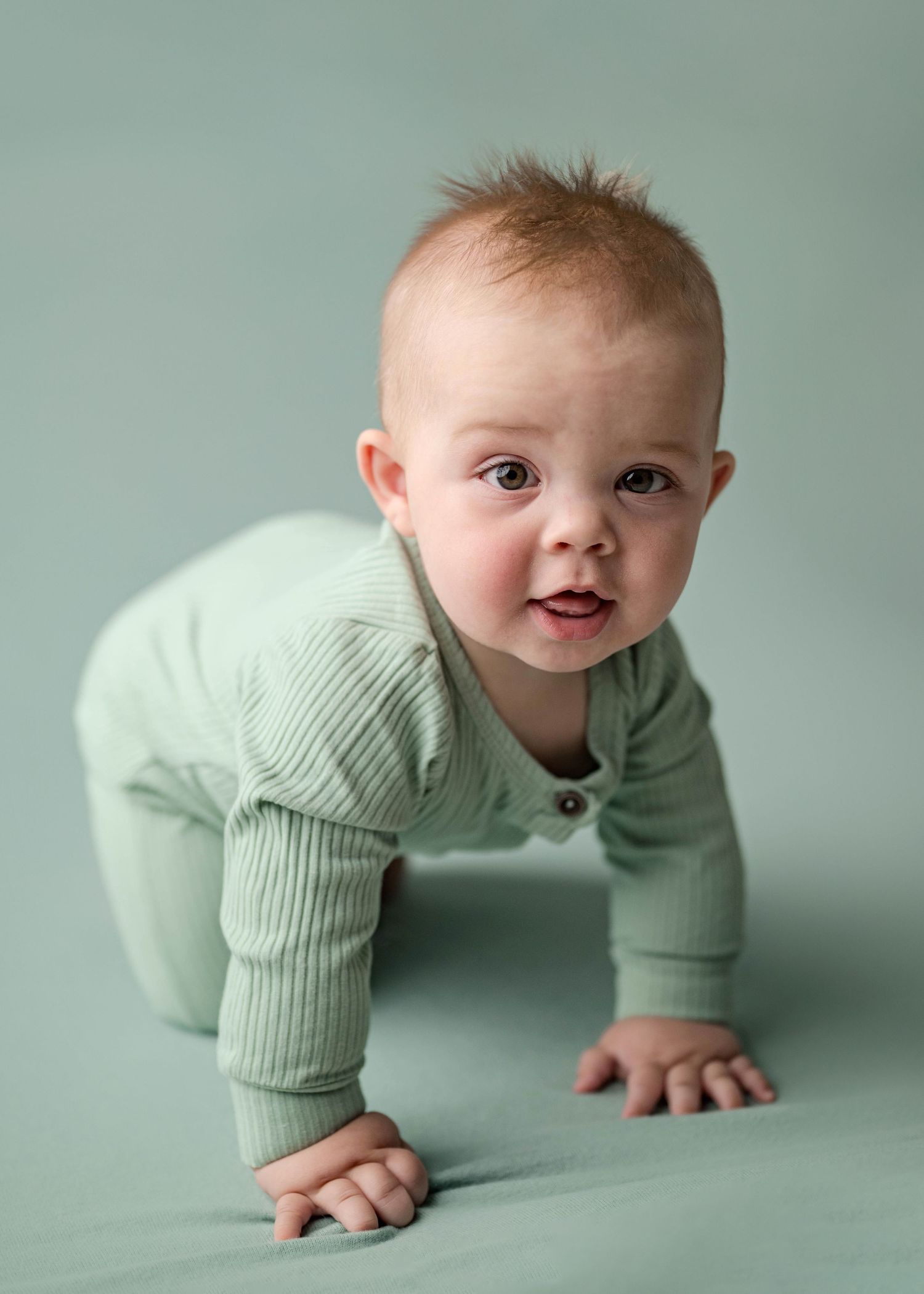 Cute red-haired baby in sage green ribbed romper crawling toward camera in professional studio portrait.