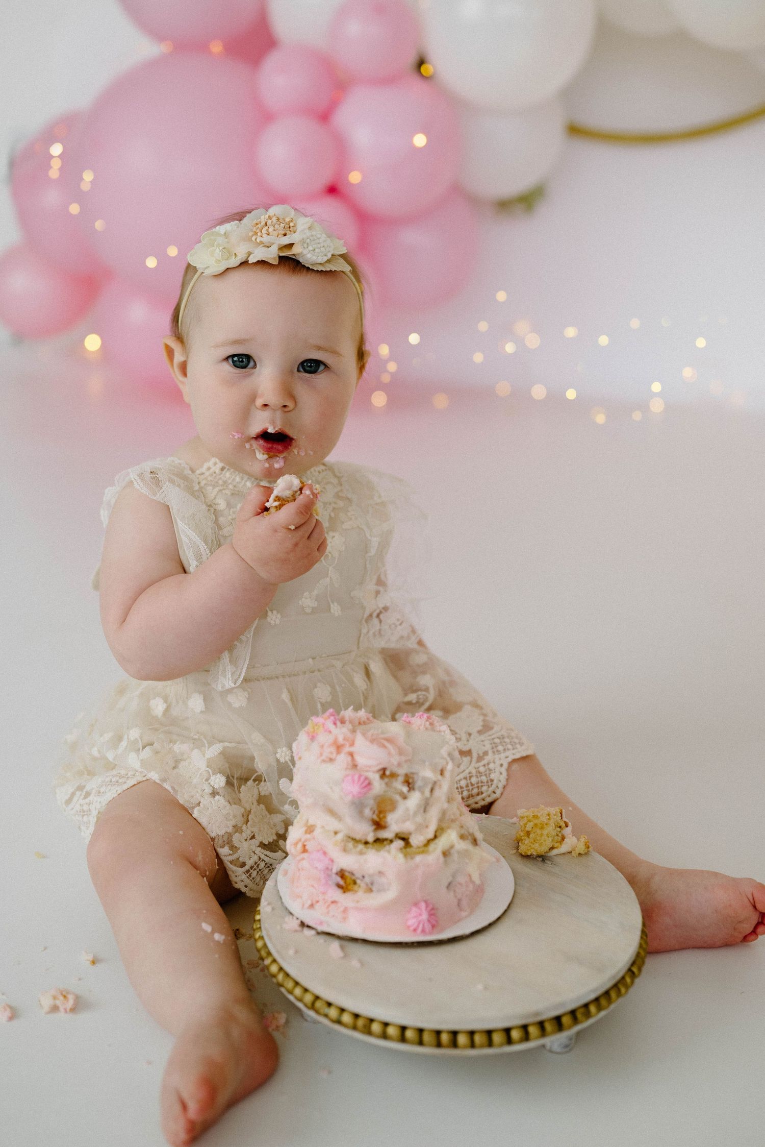 Baby girl in lace romper eats smash cake at first birthday with pink and gold balloon garland backdrop.