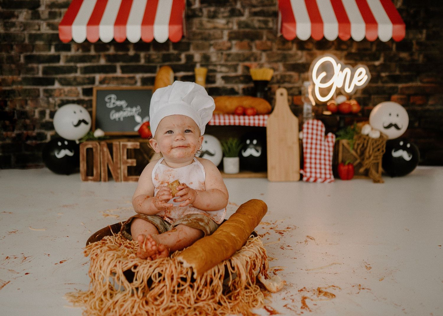 Happy baby in chef hat sits in spaghetti nest with baguette at Italian-themed first birthday shoot.