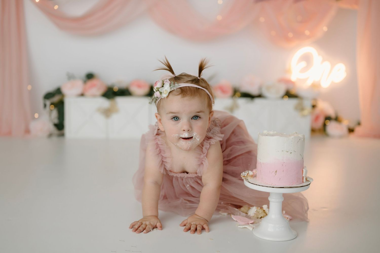 Baby girl in pink tutu dress crawls toward a pink ombre birthday cake at a floral first birthday session.