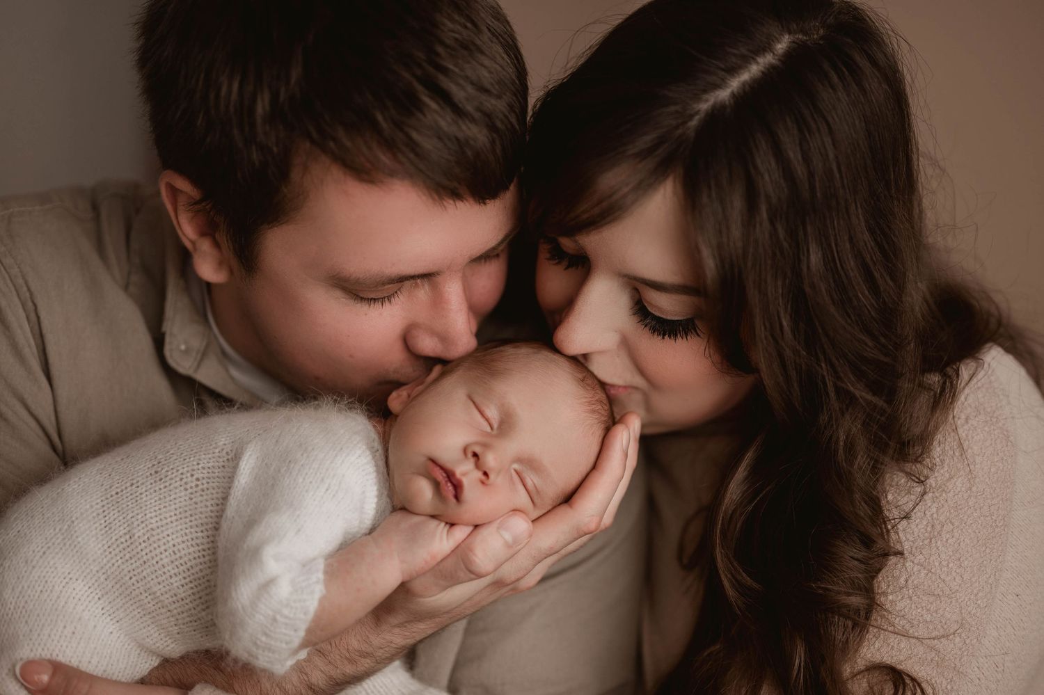 Loving parents kissing their sleeping newborn baby on the cheeks in a studio portrait.