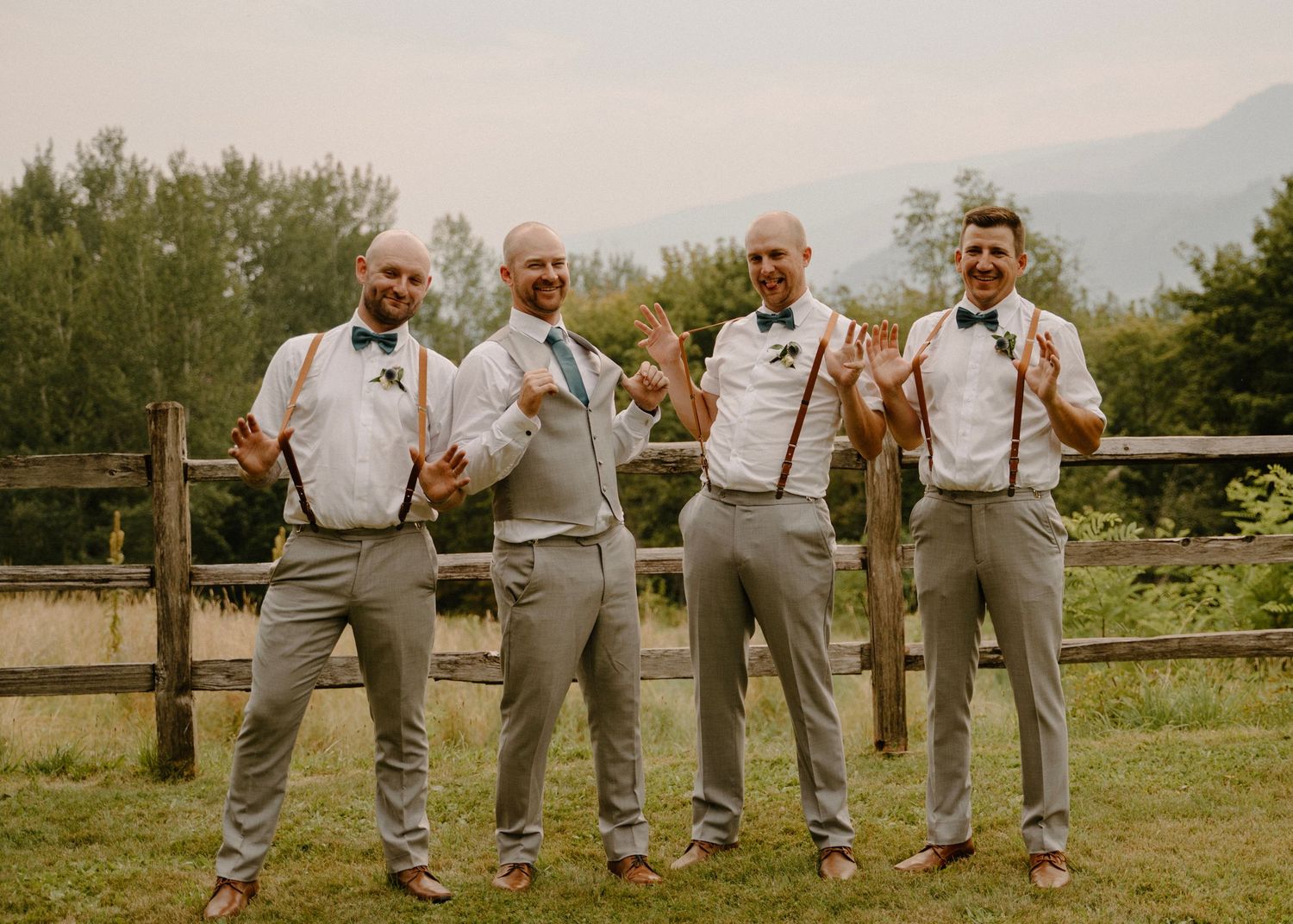 Four groomsmen in grey suits with suspenders and bow ties posing outdoors at a rustic wedding venue.