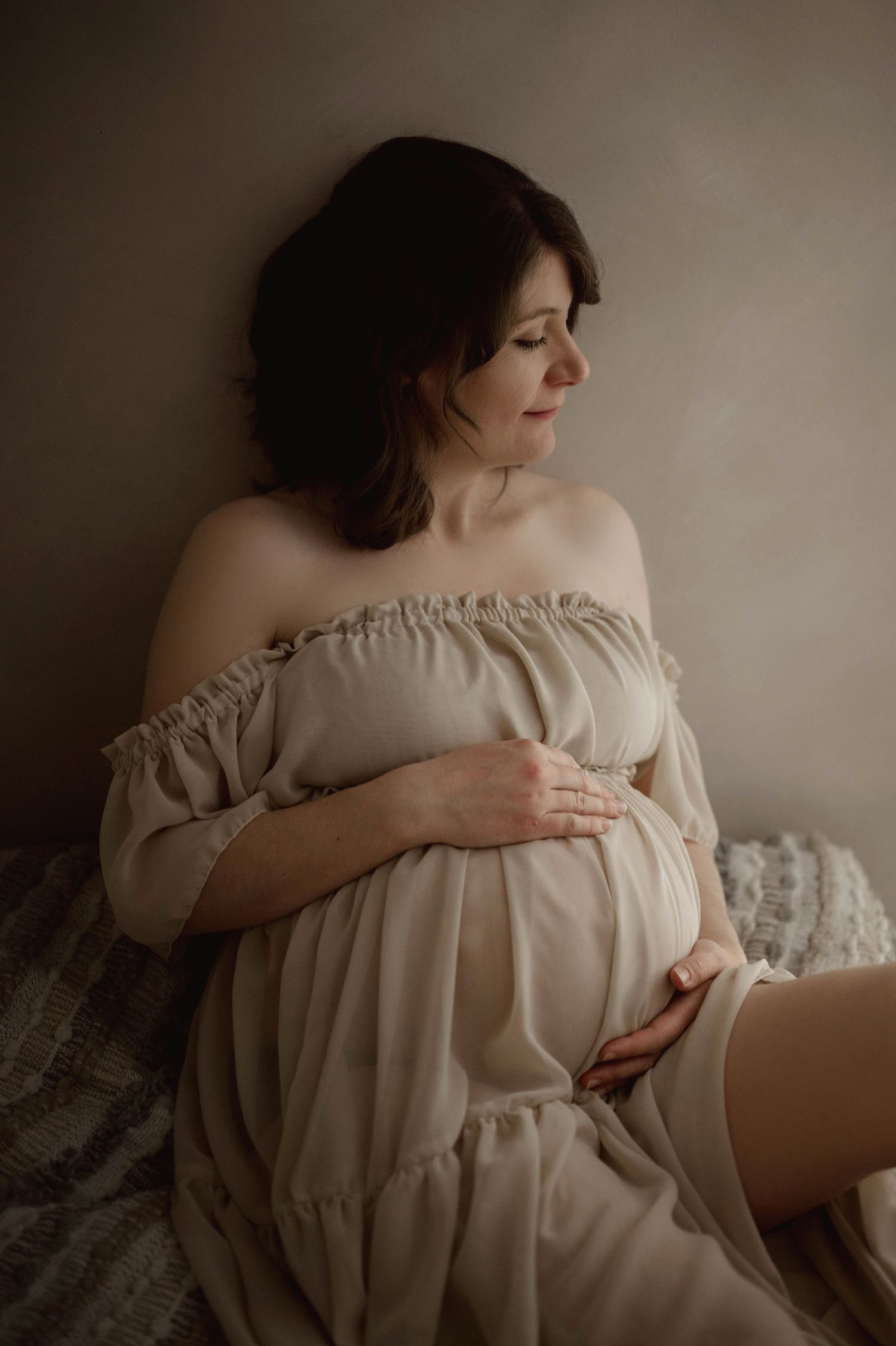 Pregnant woman in beige off-shoulder dress sitting on bed cradling bump against neutral studio backdrop.
